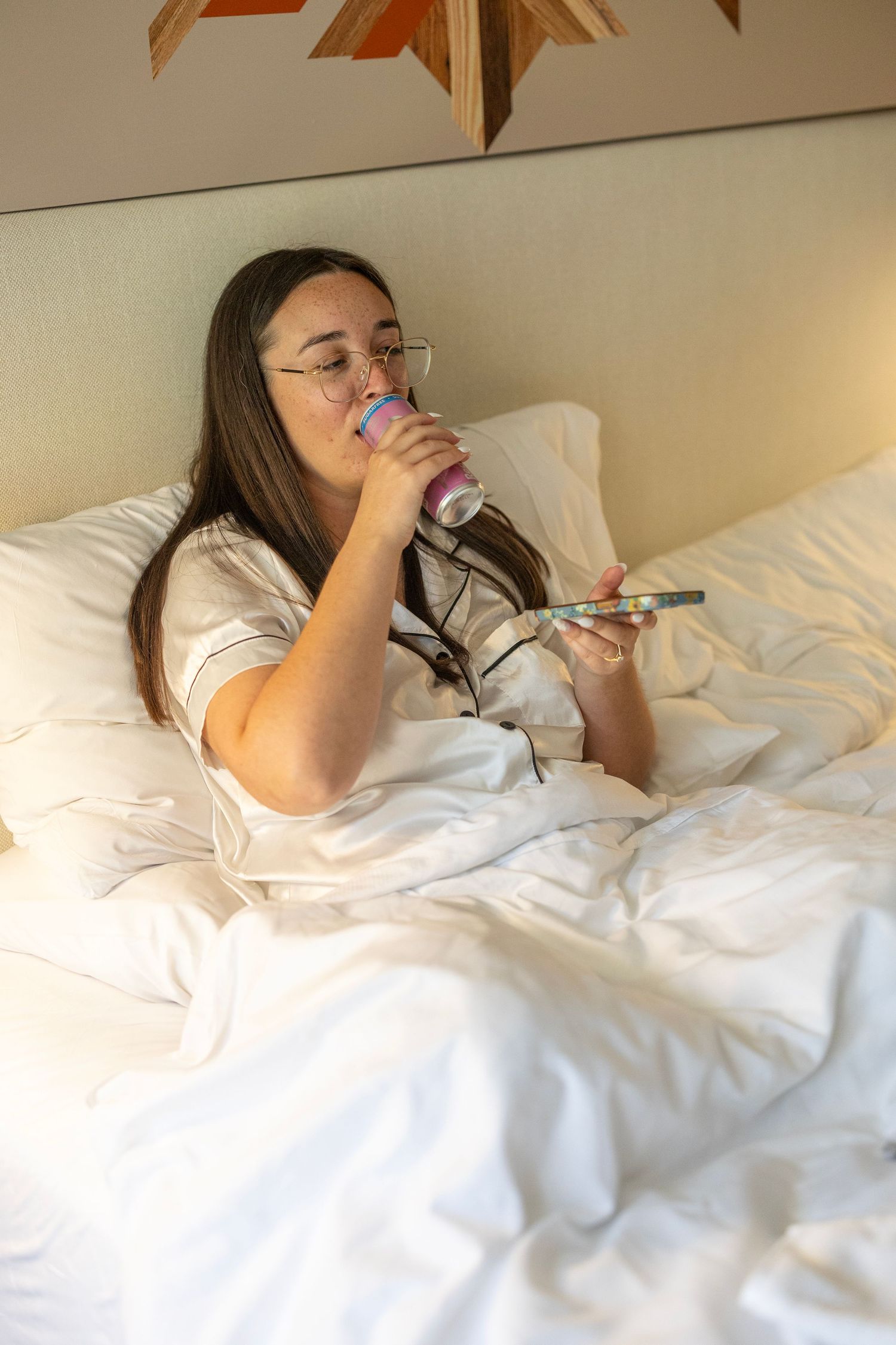 Young woman in white pajamas lying in bed, drinking from a cup while holding a TV remote control.