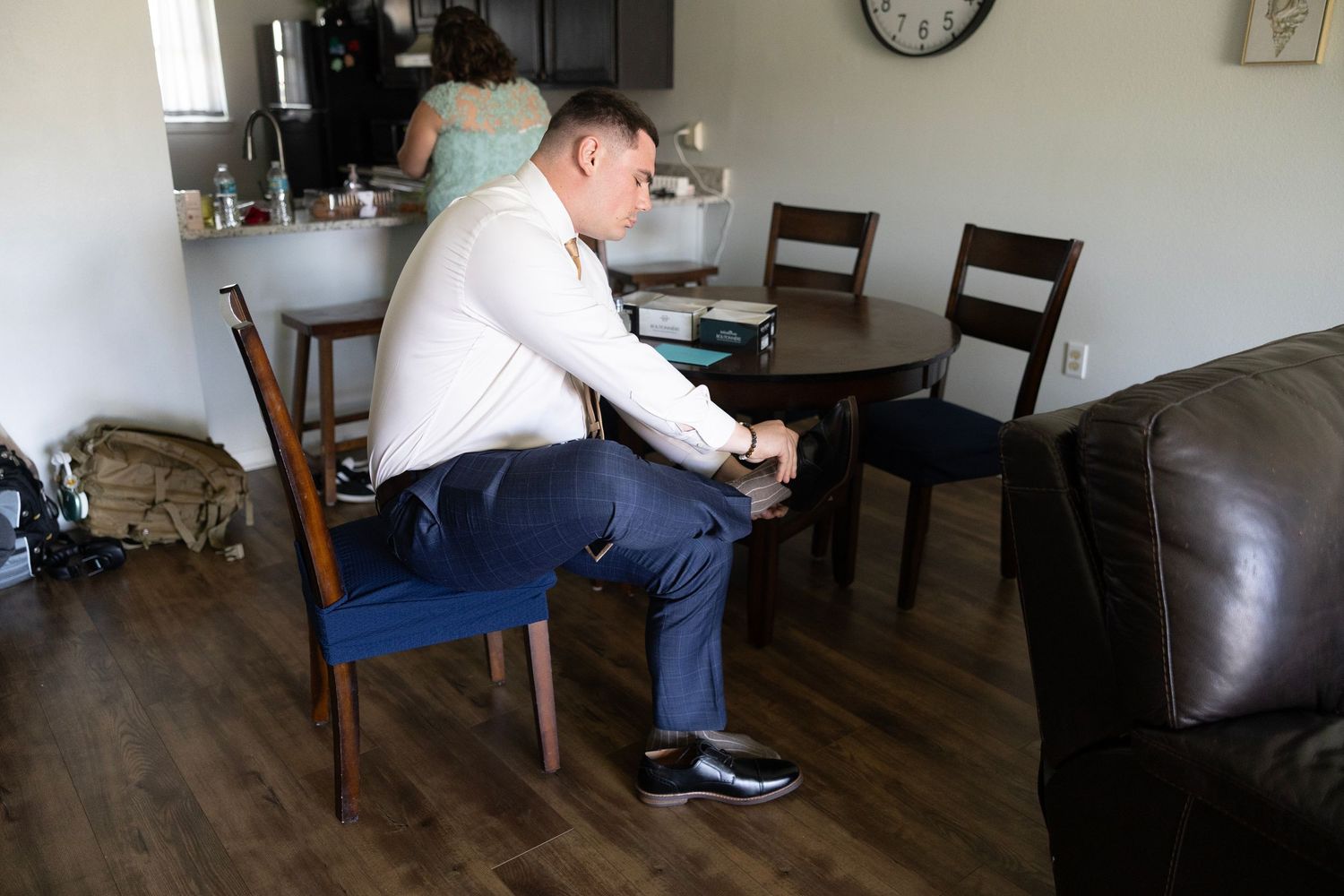 Man in white dress shirt and navy pants puts on dress shoes while sitting at dining table, woman in teal dress visible behind.