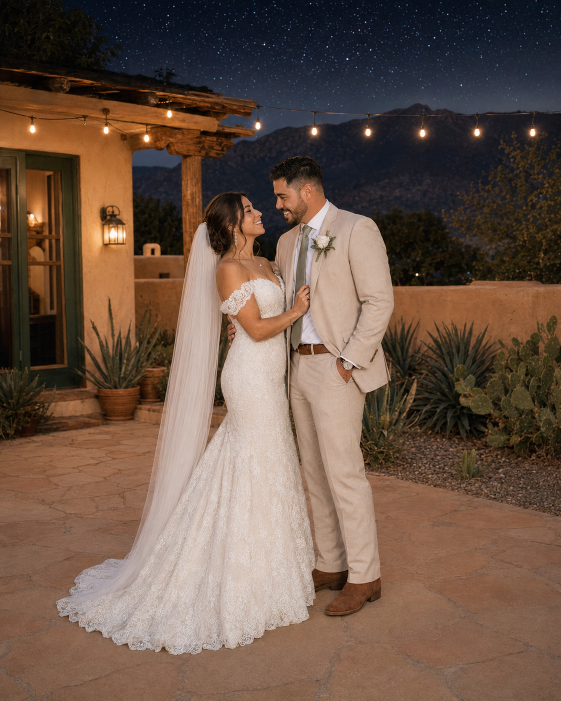 Bride in lace mermaid gown and groom in tan suit share romantic moment under starry desert sky with string lights.