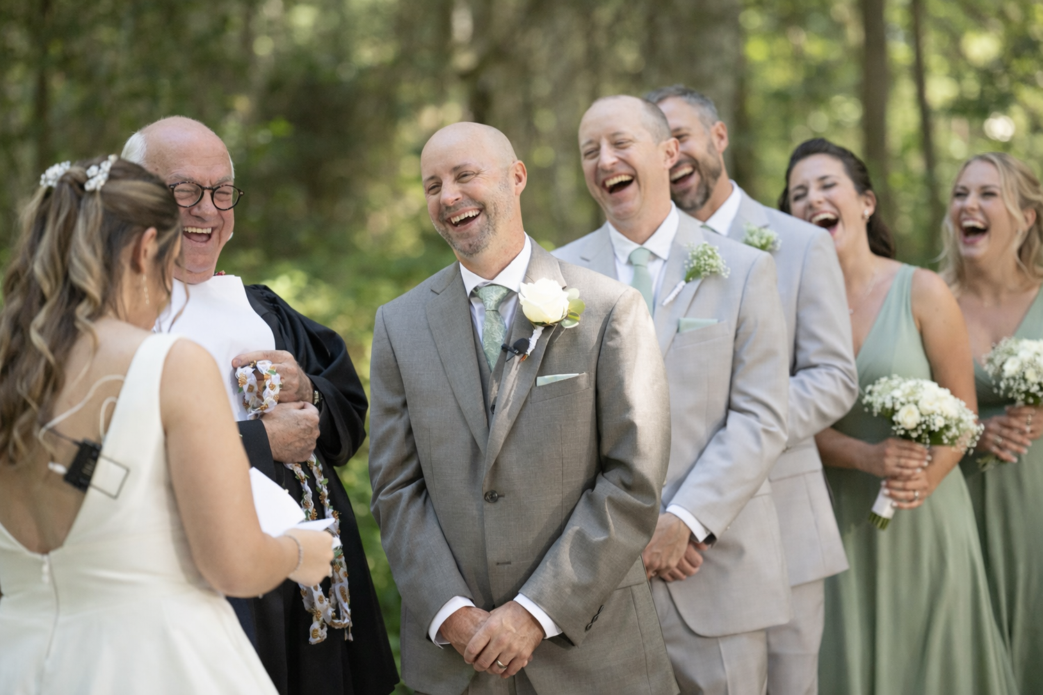 Joyful wedding party laughing outdoors in forest, groom in gray suit with white boutonniere, bridesmaids in sage green dresses.