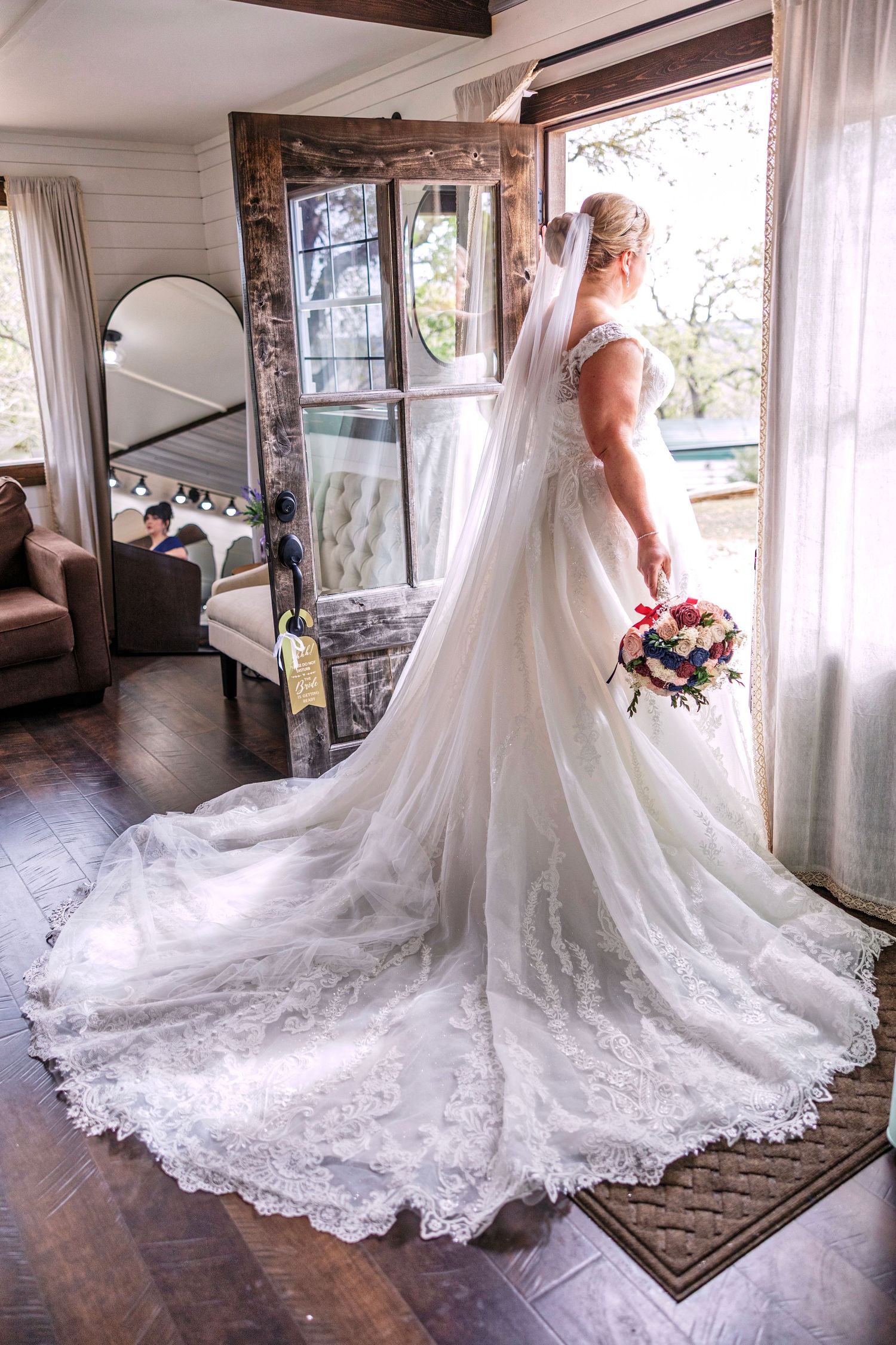 Bride in elegant lace wedding gown with long train stands by rustic door holding colorful bouquet, veil flowing gracefully.