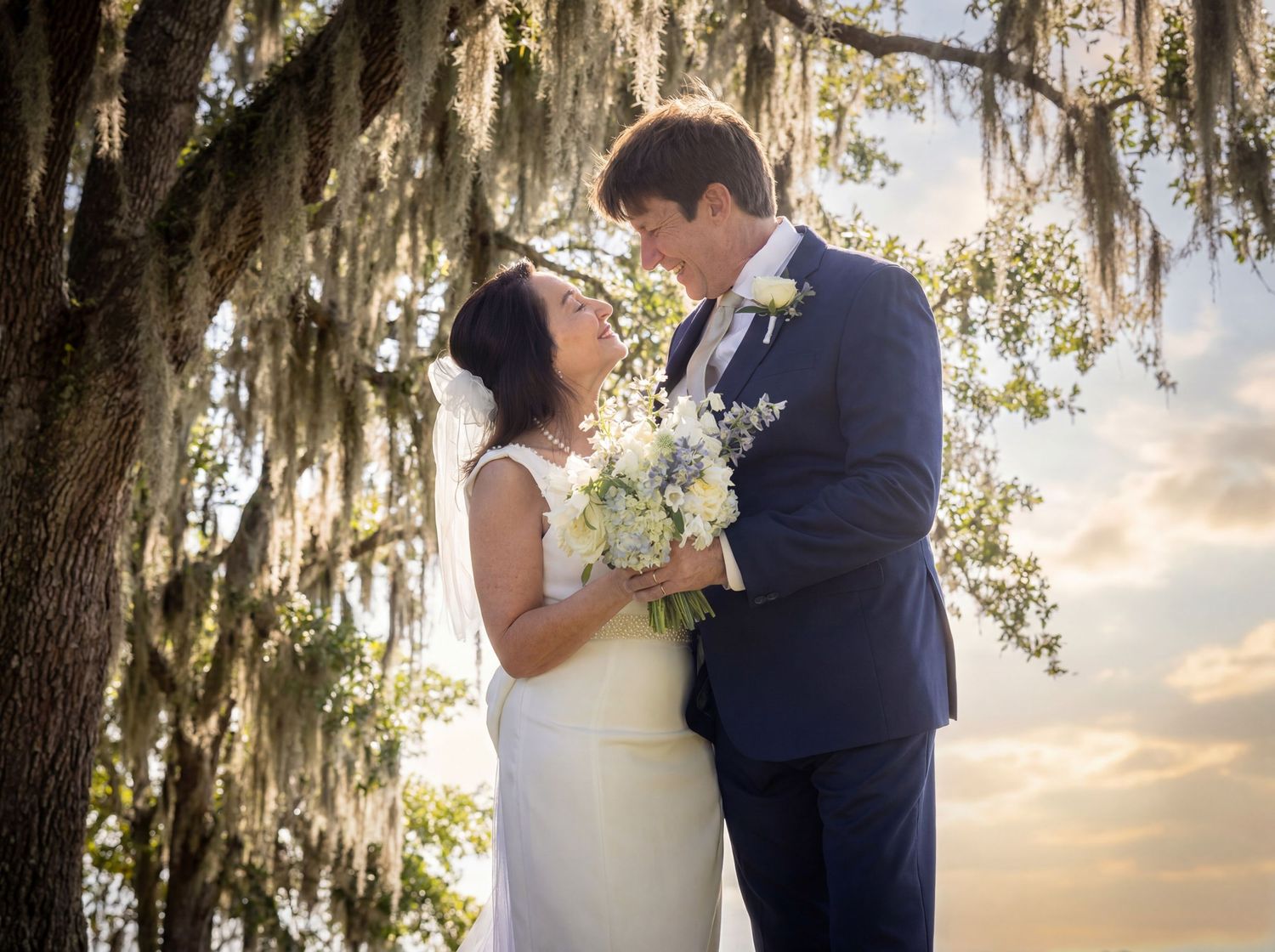 Bride and groom share a romantic moment under Spanish moss-draped oak tree at sunset wedding.