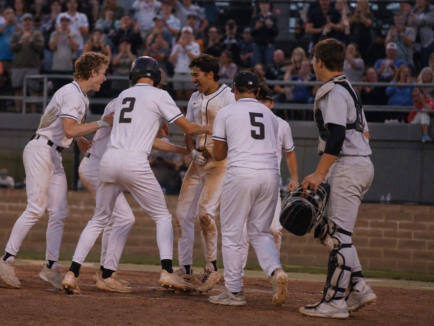 Baseball players in white uniforms celebrate at home plate after a scoring play during a night game.