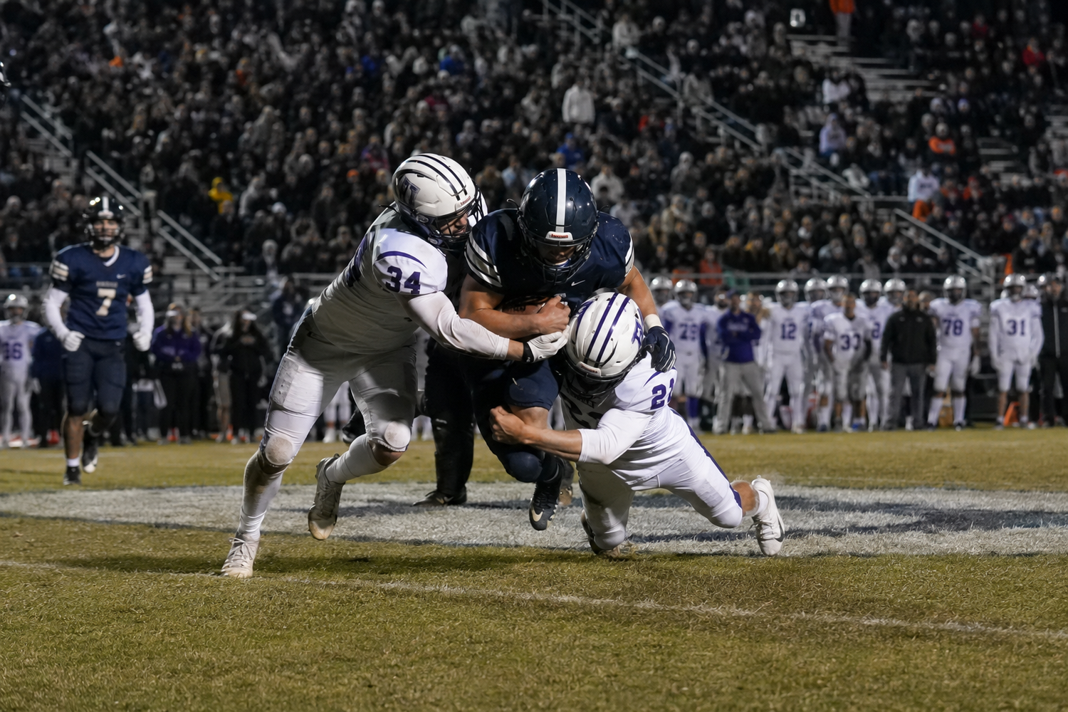 Football players tackle an opponent during a night game, with teammates and fans visible in the background.