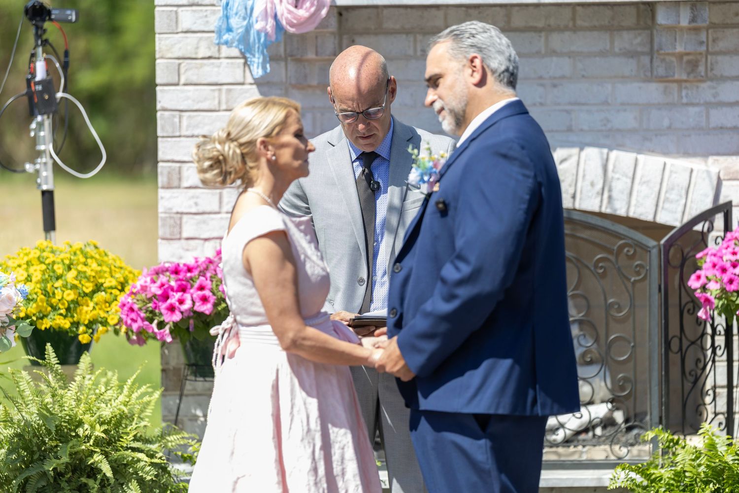 Outdoor wedding ceremony with bride and groom exchanging vows before officiant, surrounded by colorful flowers.