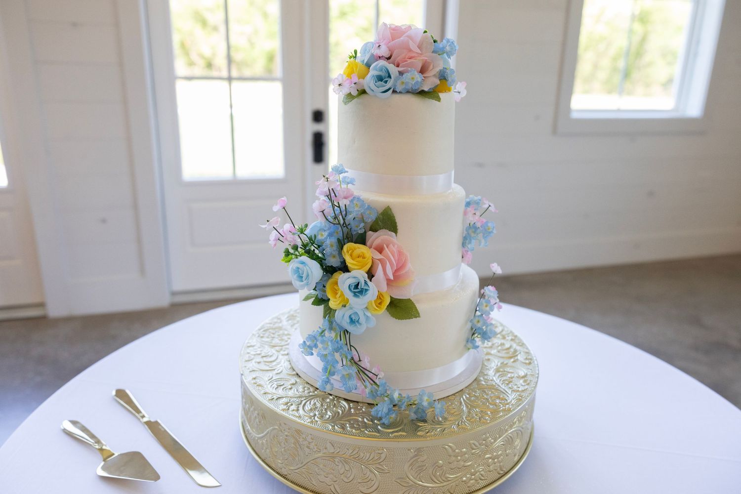 Three-tier white wedding cake adorned with colorful pink, blue, and yellow sugar flowers on a gold stand.