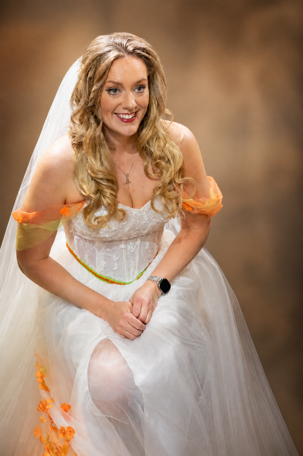 Smiling bride in white and orange wedding gown with veil and smartwatch poses elegantly for a portrait photo.