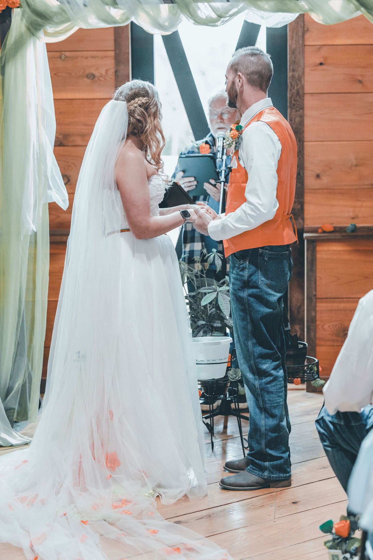 Bride and groom holding hands during indoor wedding ceremony, officiant reading vows, rustic wood barn venue.
