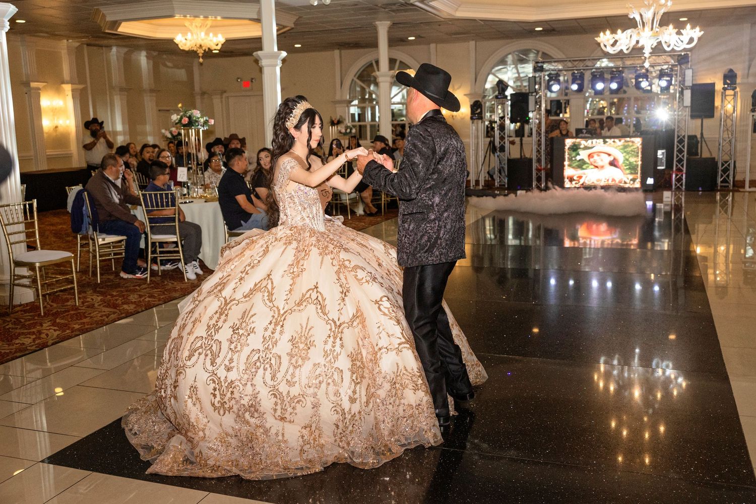 A young woman in a golden ballgown dances with a man in a black cowboy hat at an elegant quinceañera celebration.