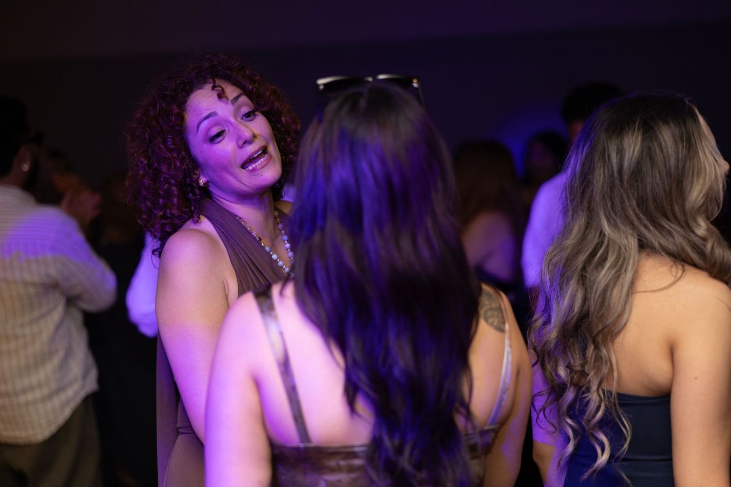 Women socializing at a nightclub party under purple lights, wearing elegant dresses in a lively crowd.