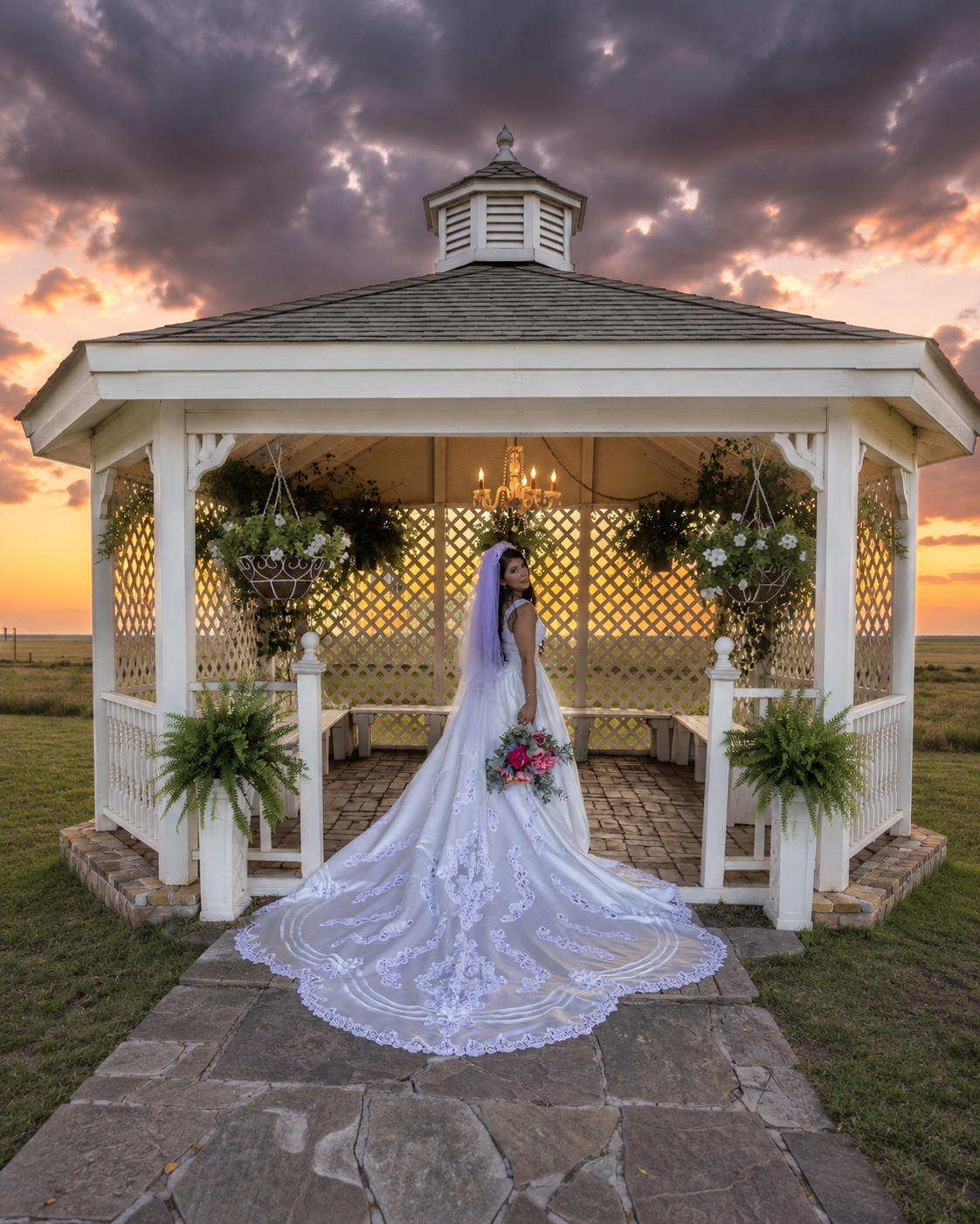 Bride in white gown and veil stands in a white gazebo at sunset with dramatic clouds and warm golden light.