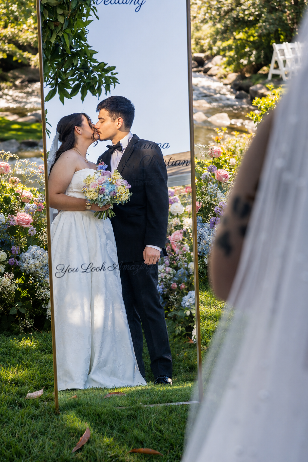 Bride and groom share a kiss reflected in a large mirror at an outdoor wedding surrounded by colorful floral arrangements.