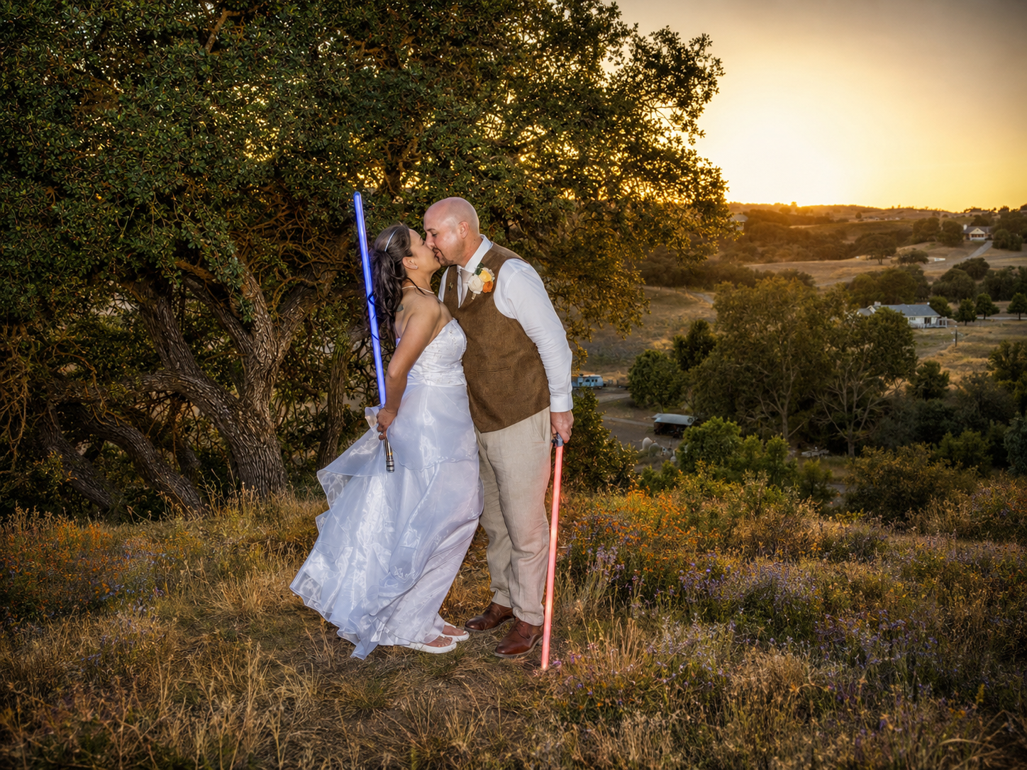 Couple sharing a sunset kiss on a hillside in wedding attire, holding blue and red lightsabers in a Star Wars themed ceremony.