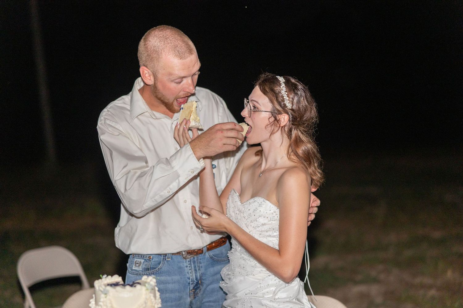 Bride and groom sharing wedding cake at outdoor nighttime reception, groom feeding cake to bride in white strapless gown.