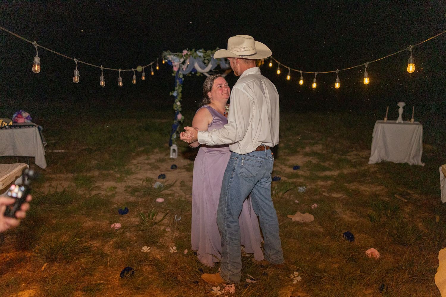 Couple dancing outdoors at night under string lights and floral arch at a rustic country wedding.