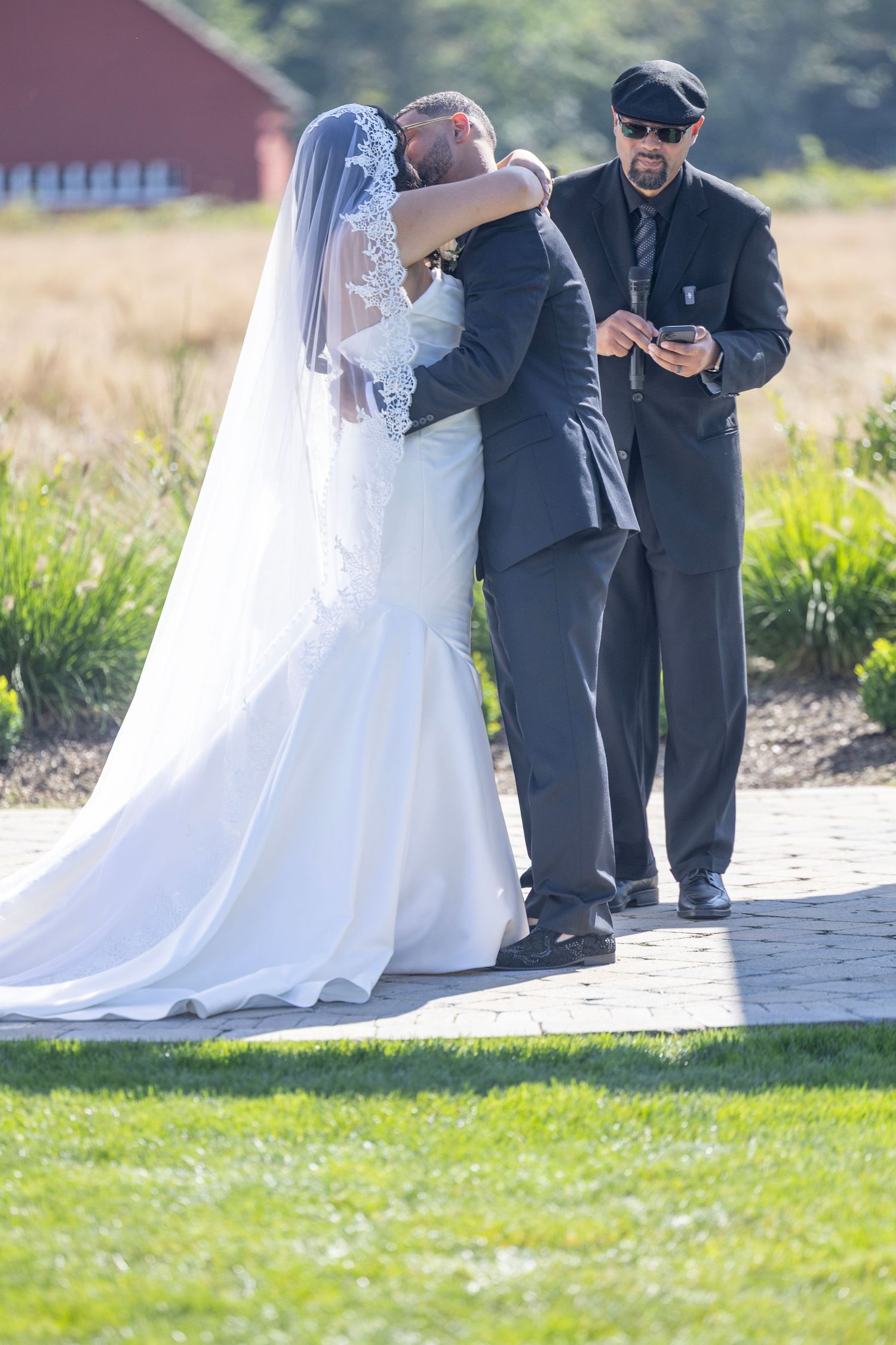 A wedding ceremony takes place outdoors with a bride in white dress and officiant in black against a rural backdrop.
