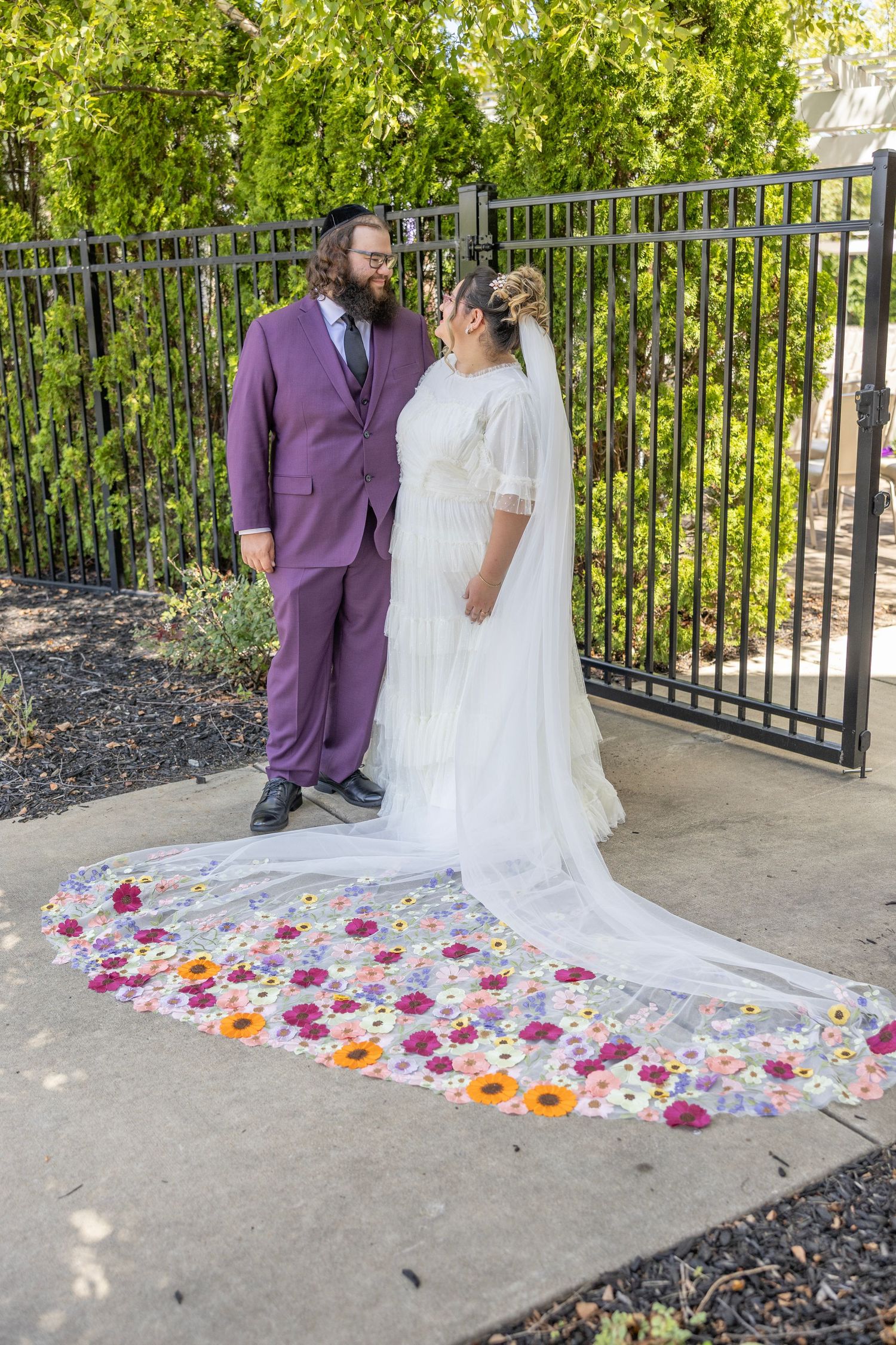 Wedding couple stands by iron fence with colorful flower petals scattered along white dress train on concrete path.