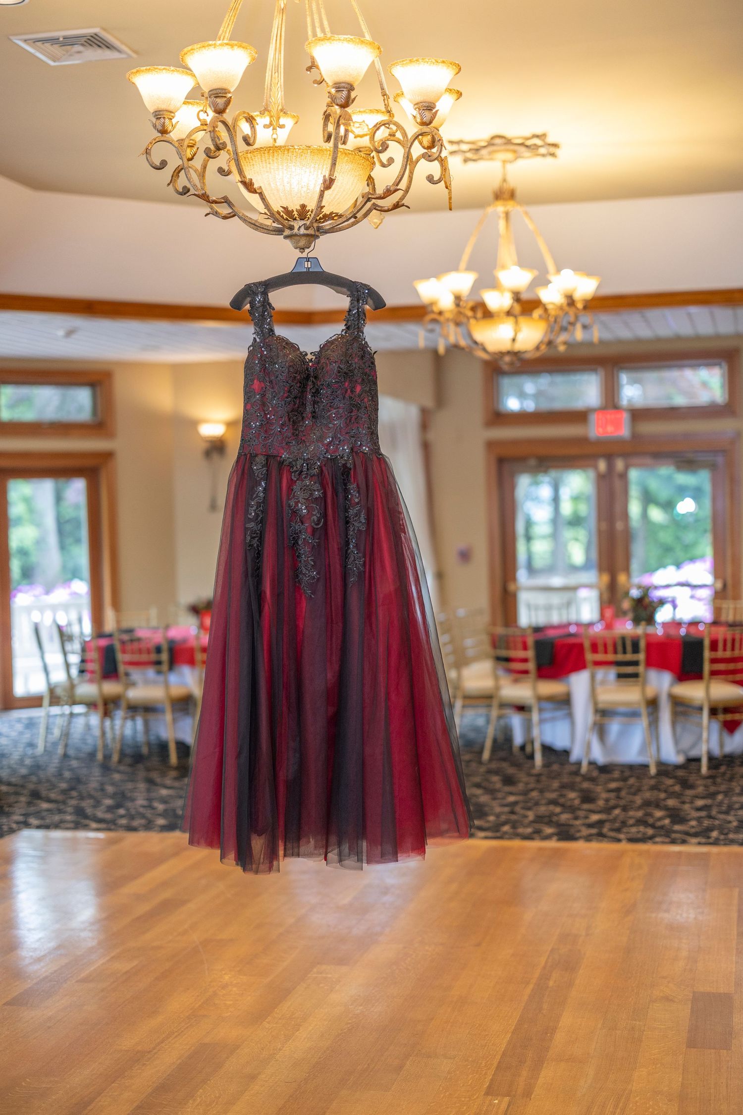 A red and black tulle dress hangs from a chandelier in an elegant ballroom with hardwood floors and warm lighting.