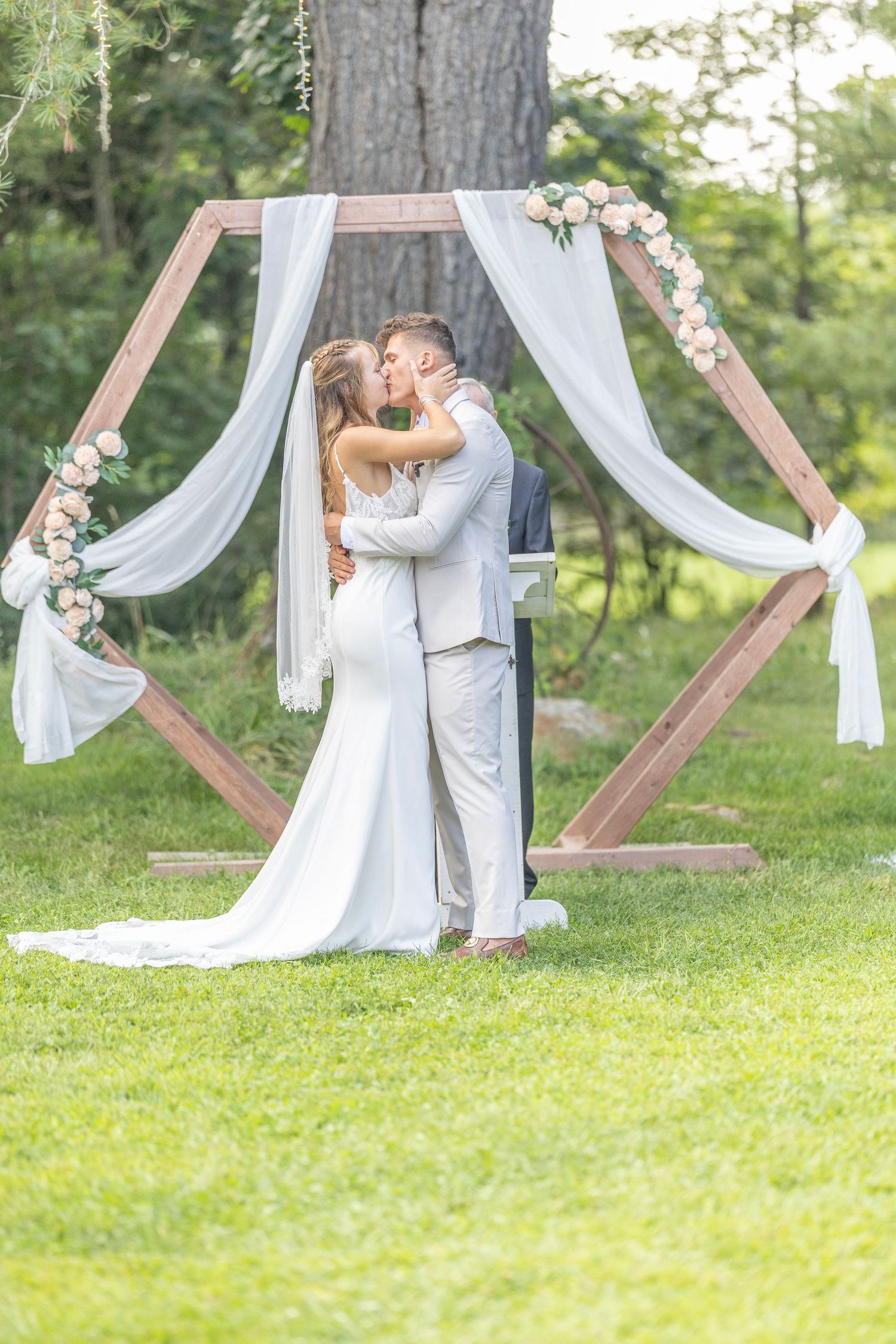 Romantic outdoor wedding ceremony with wooden triangular arch and flowing white fabric in a green garden setting.