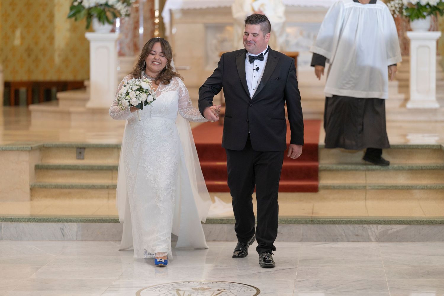 A couple walks together in formal wedding attire down a church aisle decorated with floral arrangements.