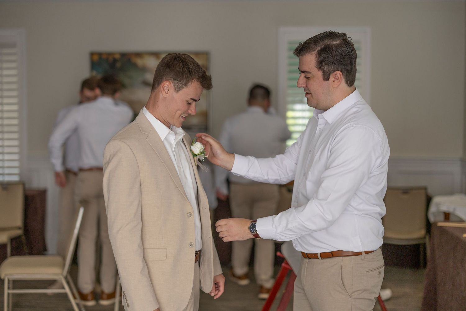 Groomsmen in tan suits and white shirts help each other get ready before a wedding ceremony.