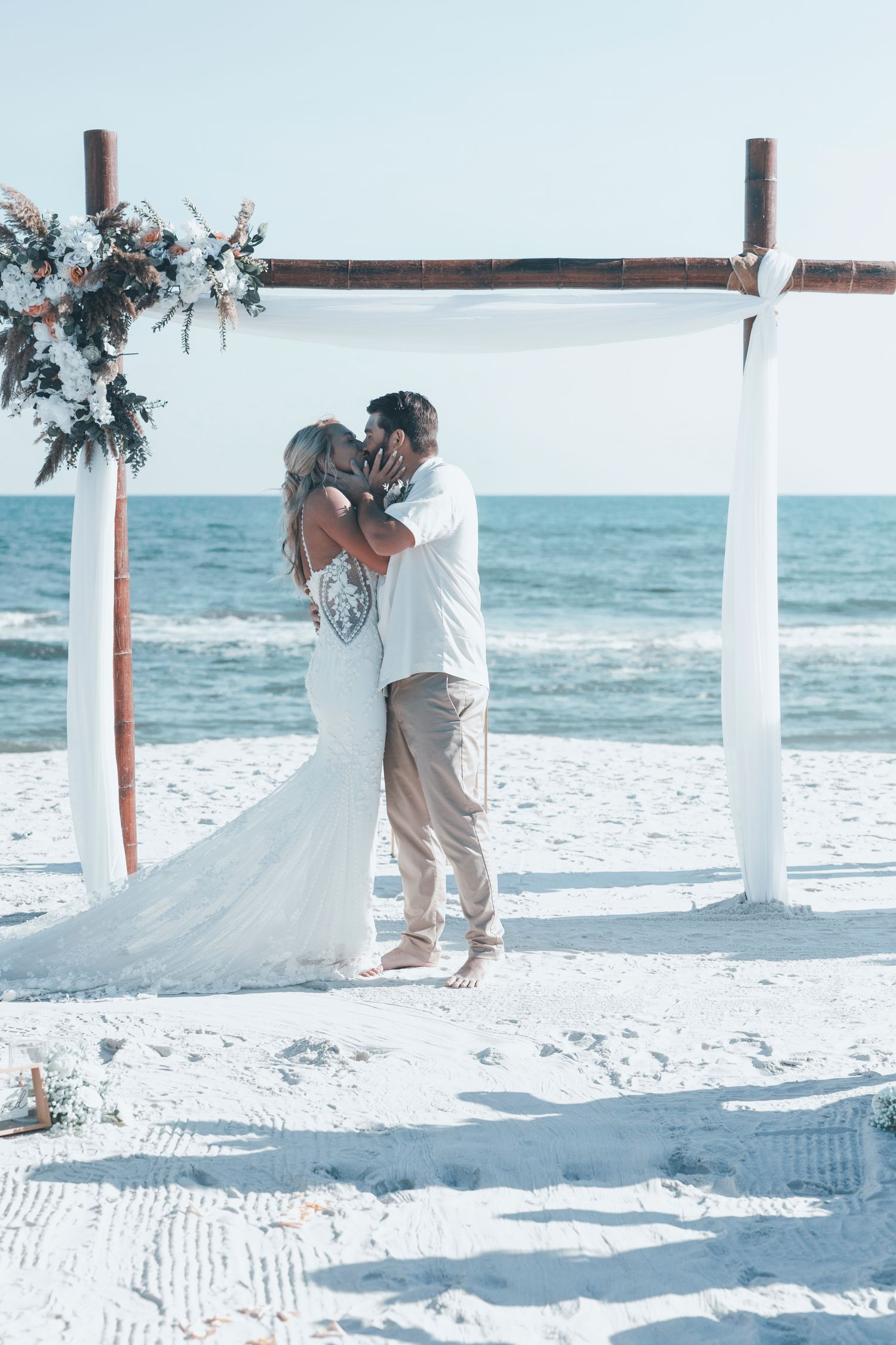 A romantic beach wedding scene with a couple embracing on pristine white sand under a wooden arch by turquoise waters.