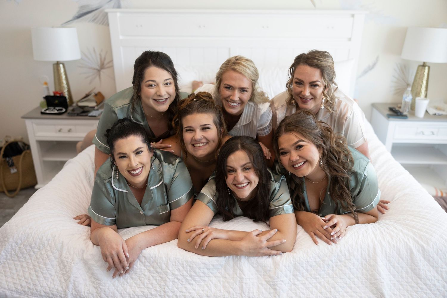 A group of friends wearing matching blue shirts smile together while lying on a white bed.