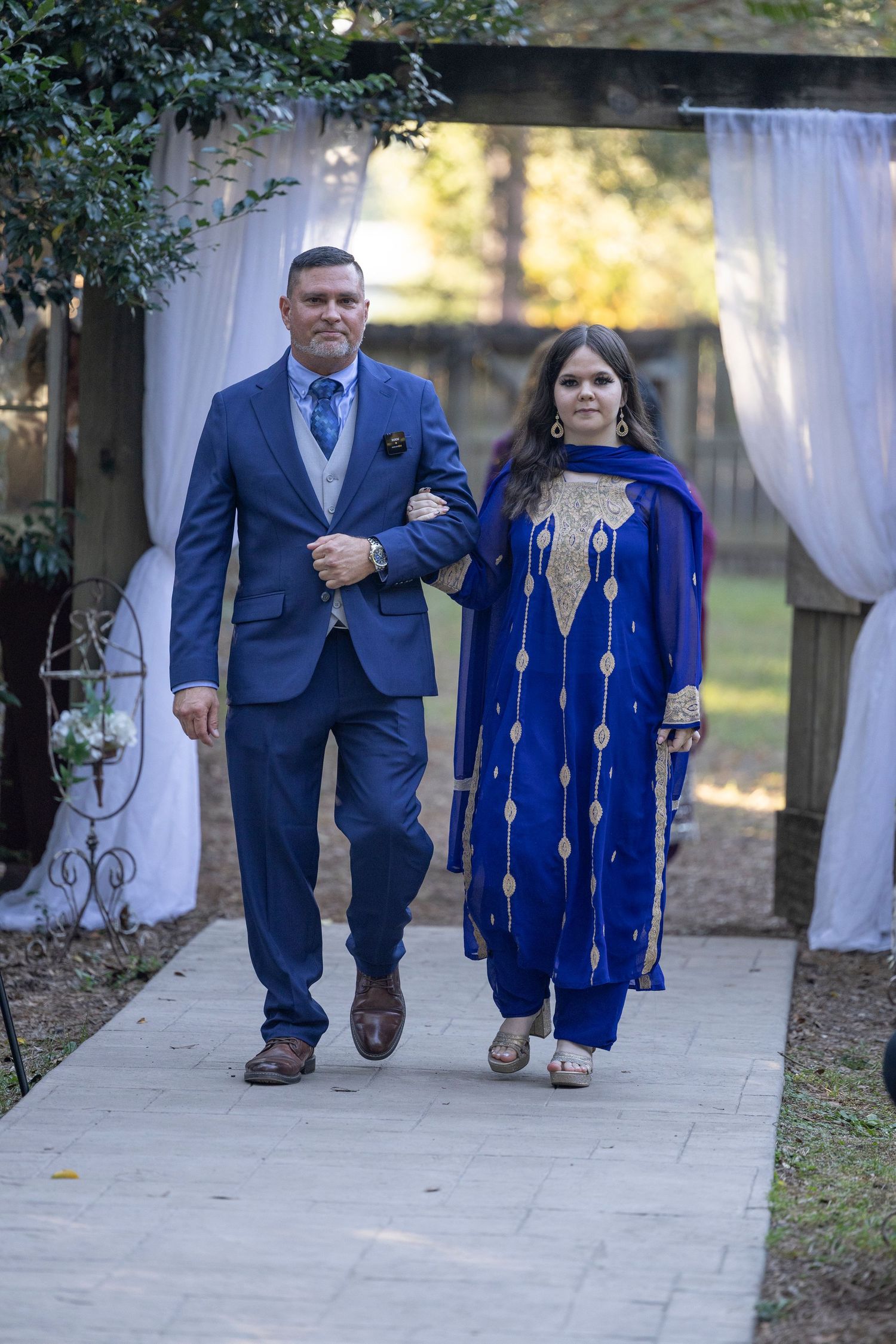 A couple in coordinating navy blue and royal blue formal attire walks together on a stone pathway at an outdoor event.