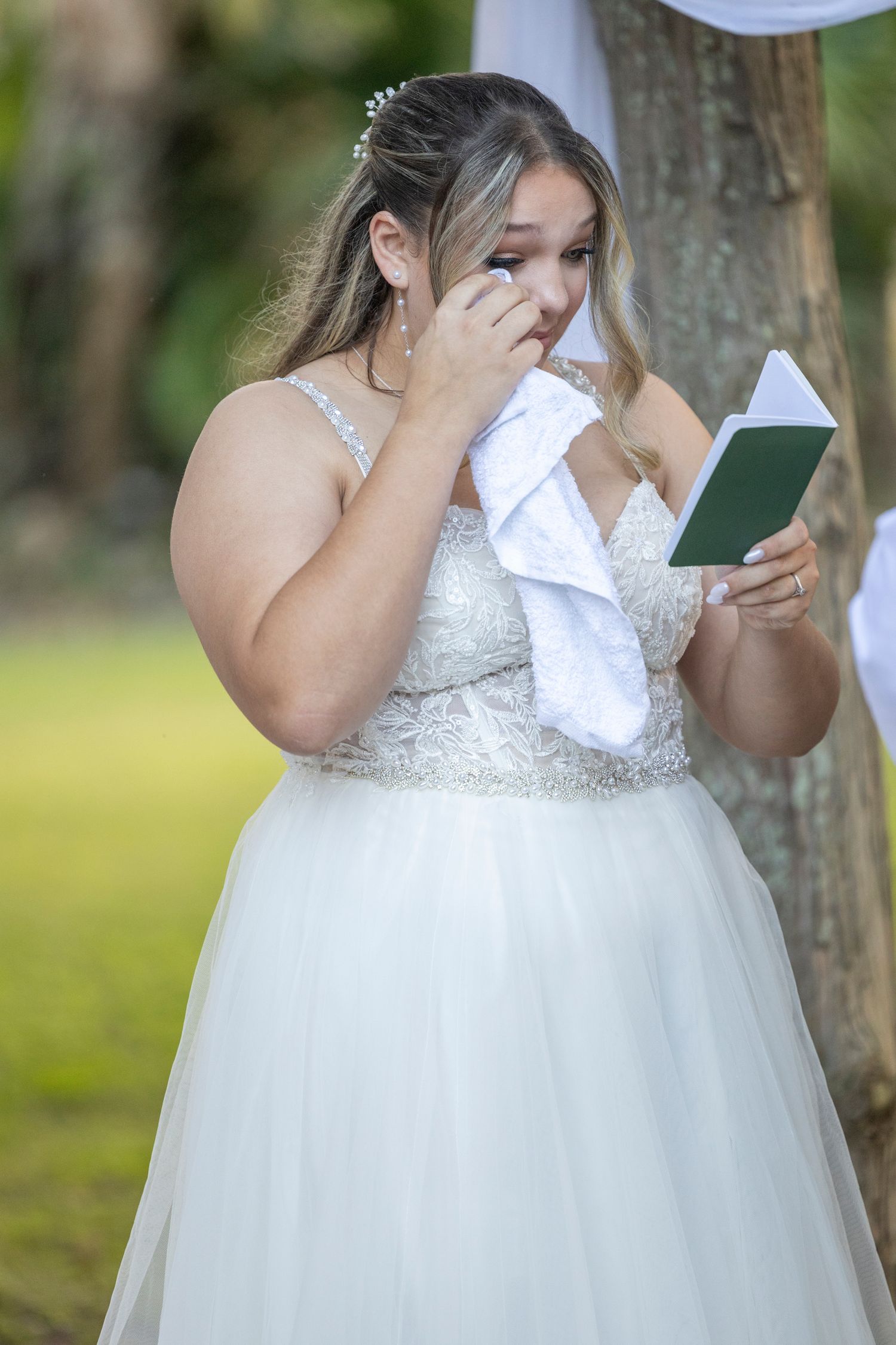 An emotional bride in a white ballgown wedding dress wipes tears with a tissue during an outdoor ceremony.