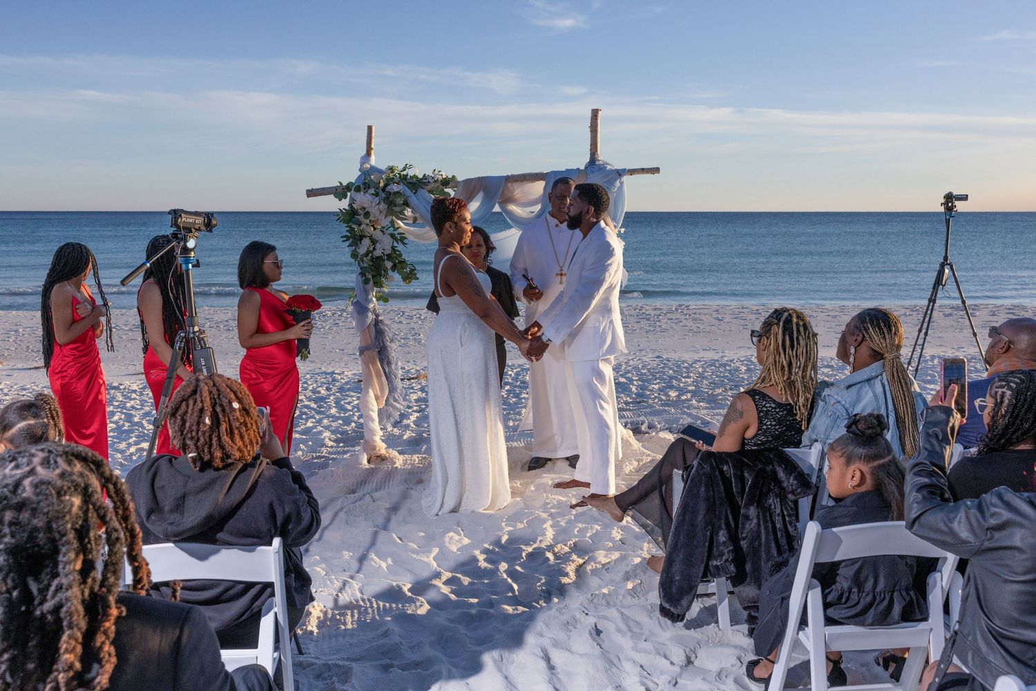 Guests gather on a sunny beach for an elegant outdoor wedding ceremony with white decor and ocean views.