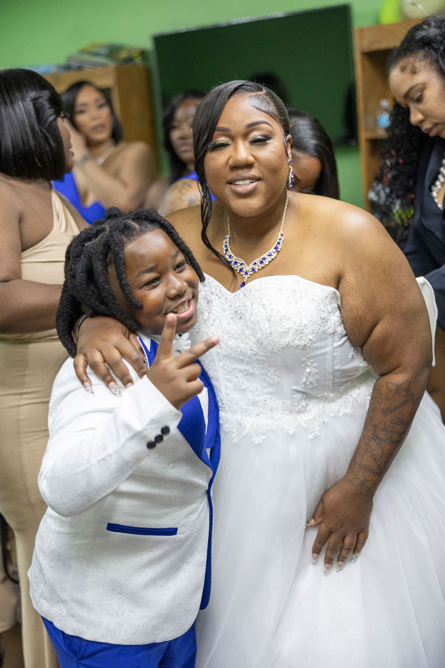 A bride in a white gown poses with a child in a white and blue suit at a wedding celebration.