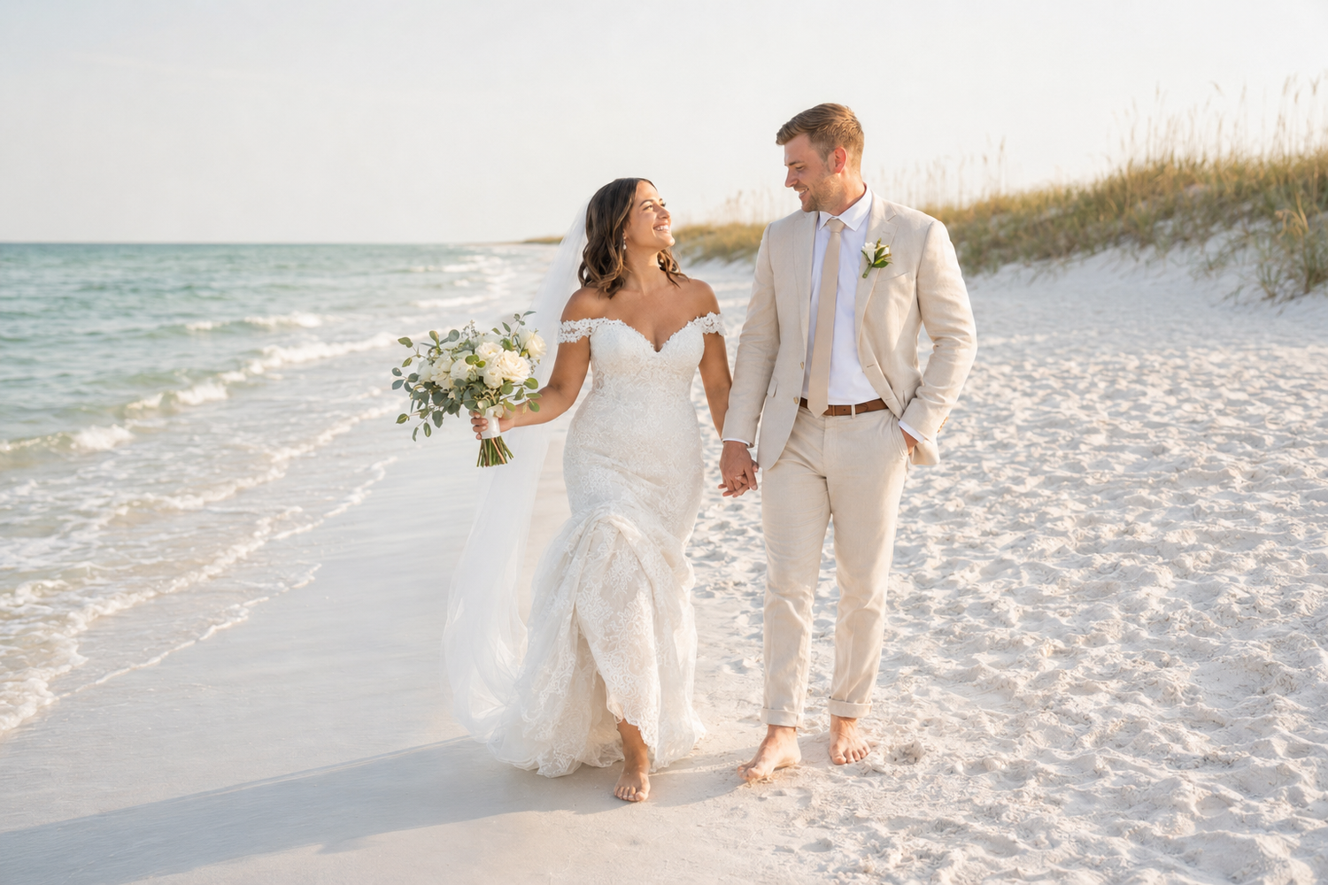 Bride and groom walk hand-in-hand on a white sand beach, waves behind them, bride holding a white floral bouquet.