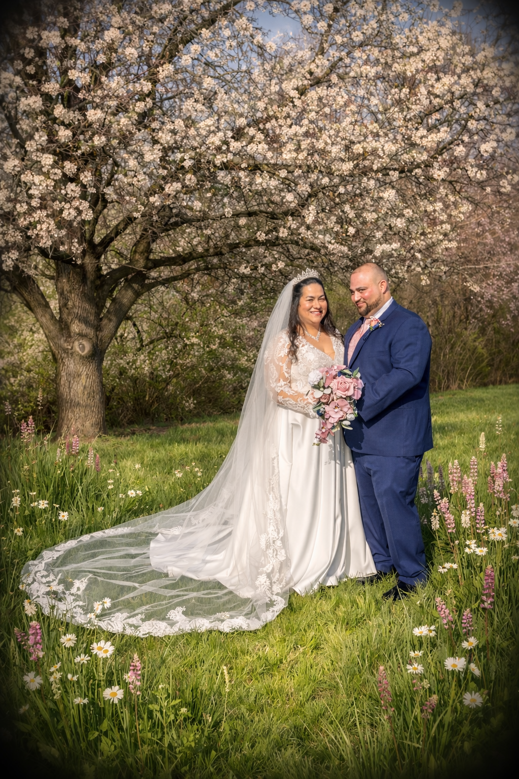 Bride and groom pose under blooming cherry blossom tree in lush meadow with wildflowers, bride in white gown with long veil.