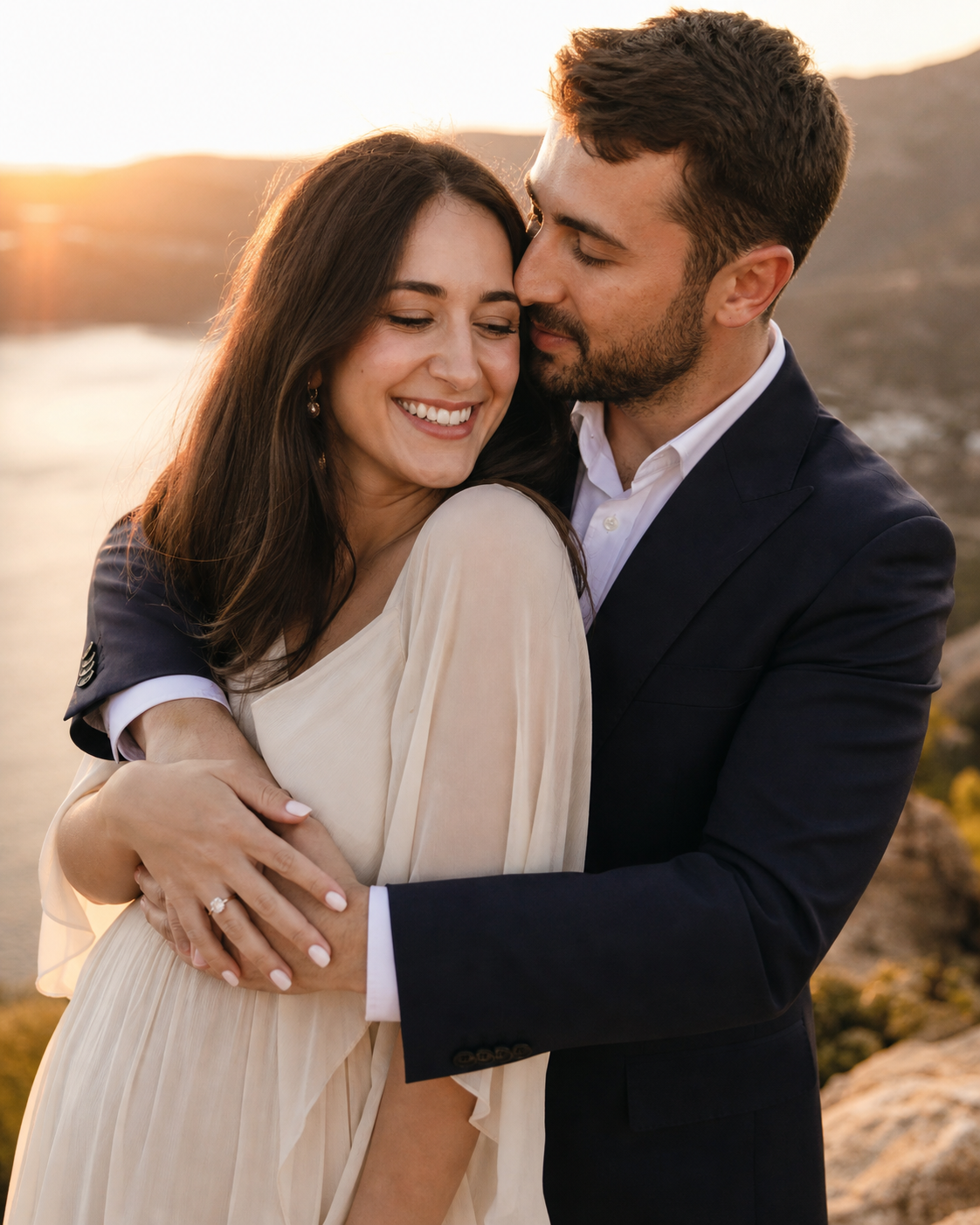 Couple embracing at golden hour sunset, woman in cream dress smiling, man in navy suit kissing her cheek outdoors.
