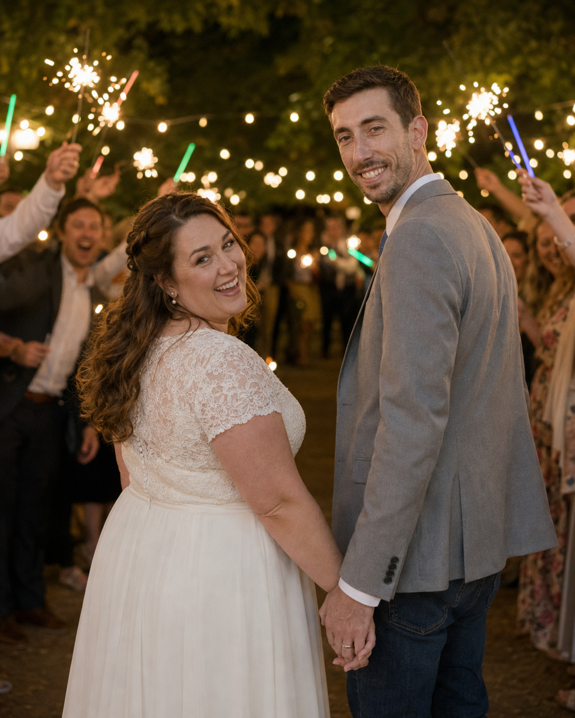 Bride and groom smile during sparkler send-off exit surrounded by cheering guests at night wedding celebration.
