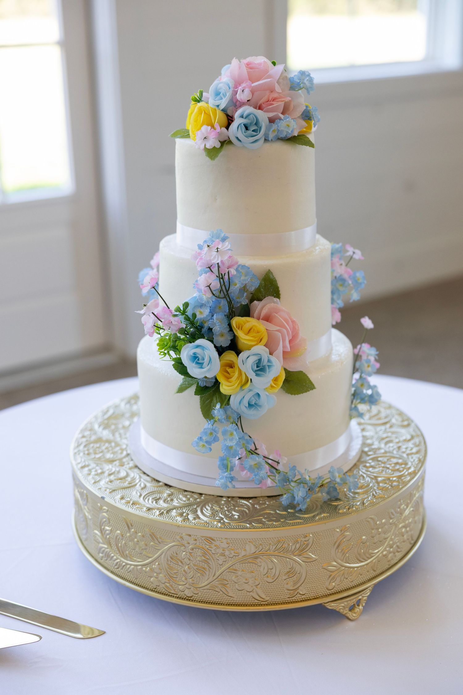 Three-tier white wedding cake with pink, blue, and yellow sugar flowers on a gold ornate cake stand.