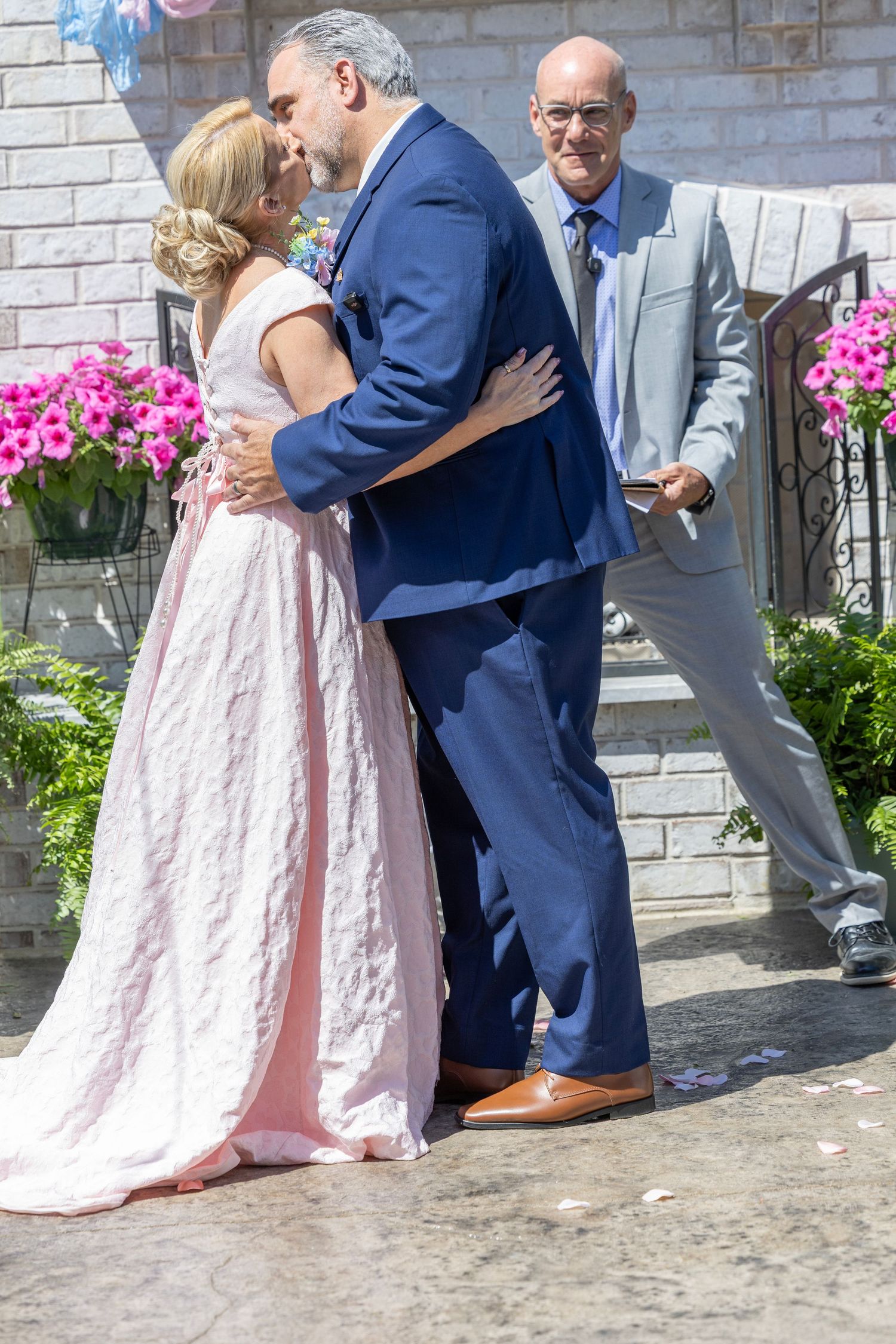 Couple sharing first kiss in wedding ceremony outdoors, bride in pink gown, officiant in gray suit watching nearby.