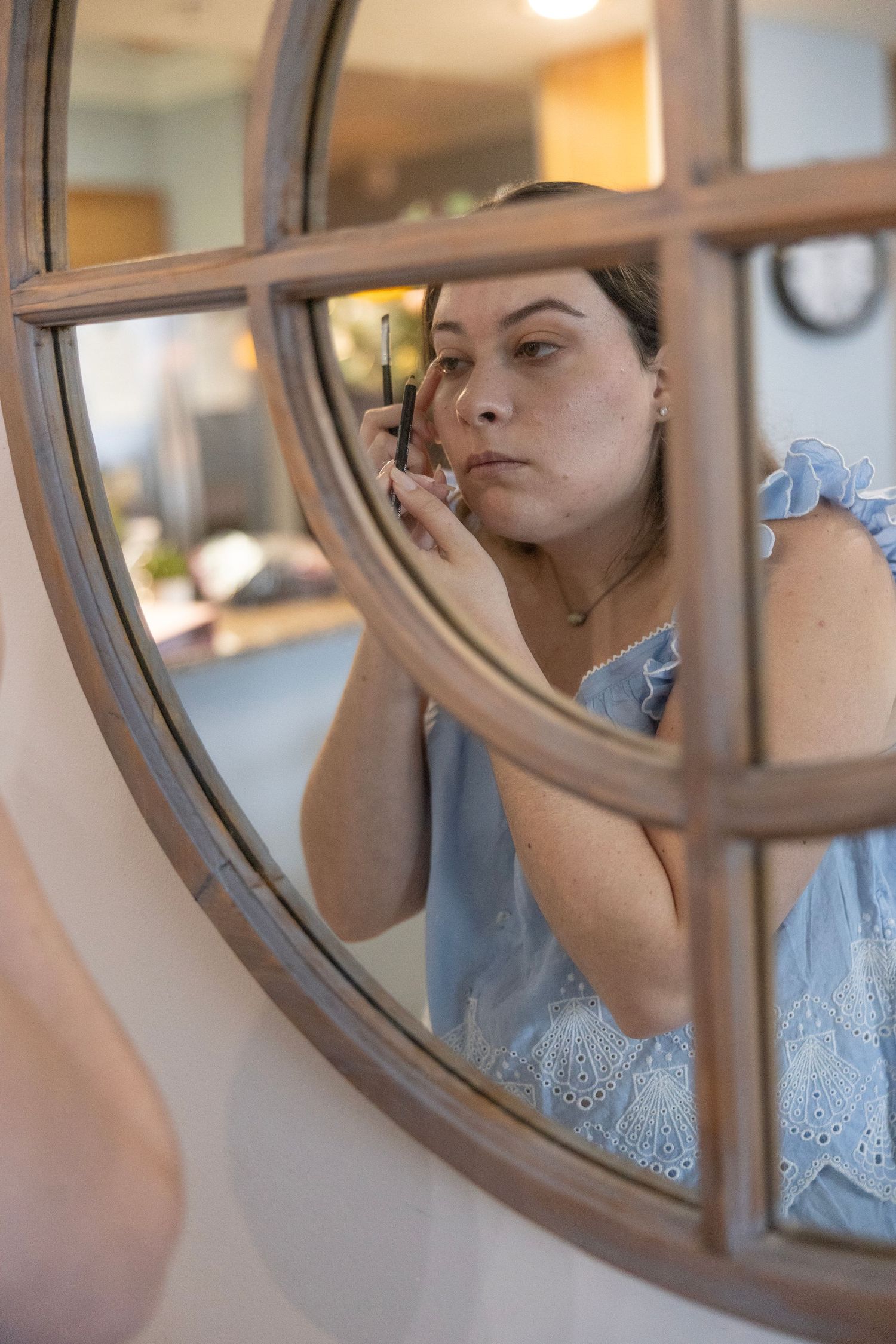 Woman applying makeup with a brush, reflected in a round wooden-framed mirror in a bright room.