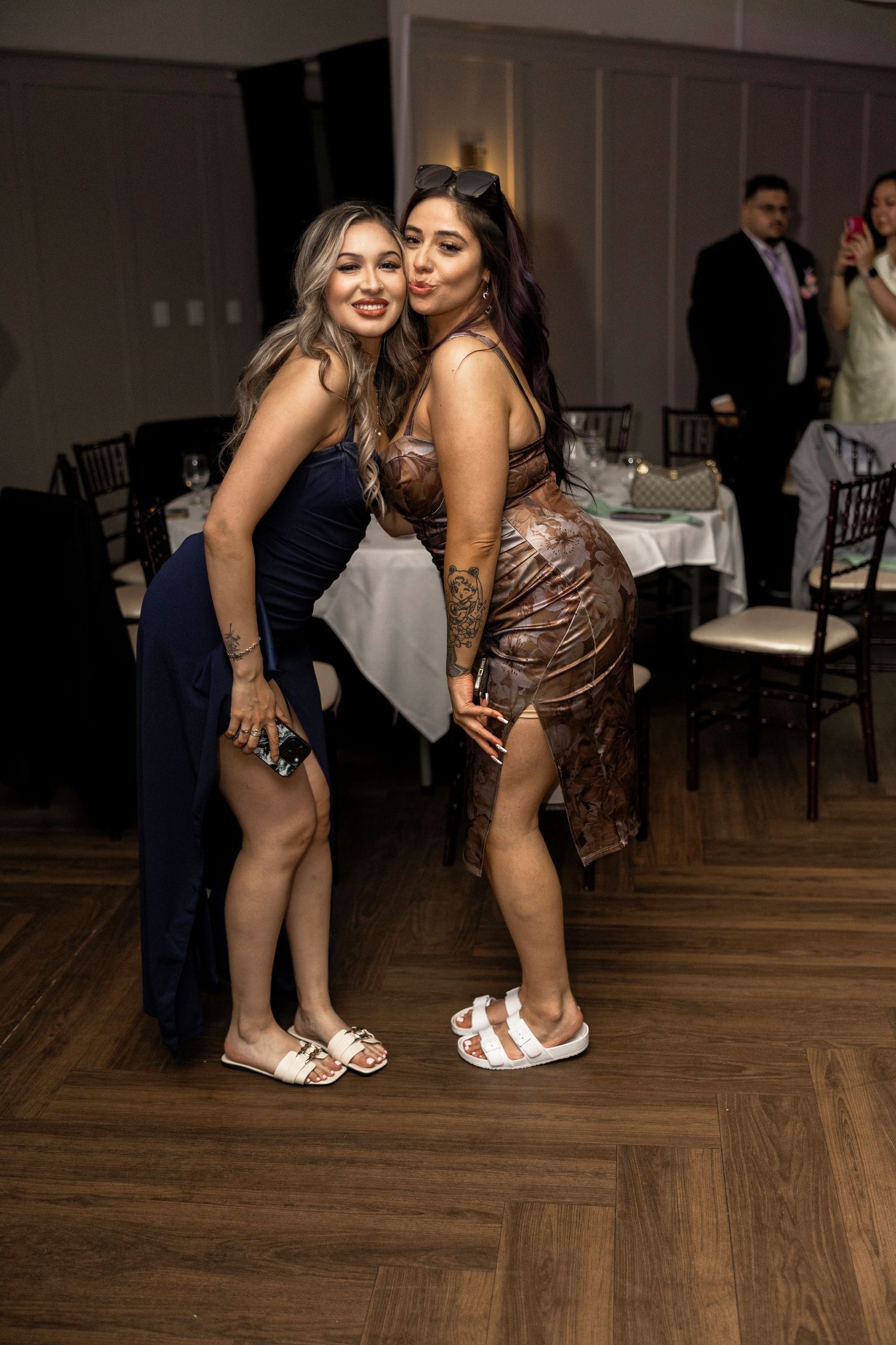Two women in elegant dresses pose together on a dance floor at a formal event.