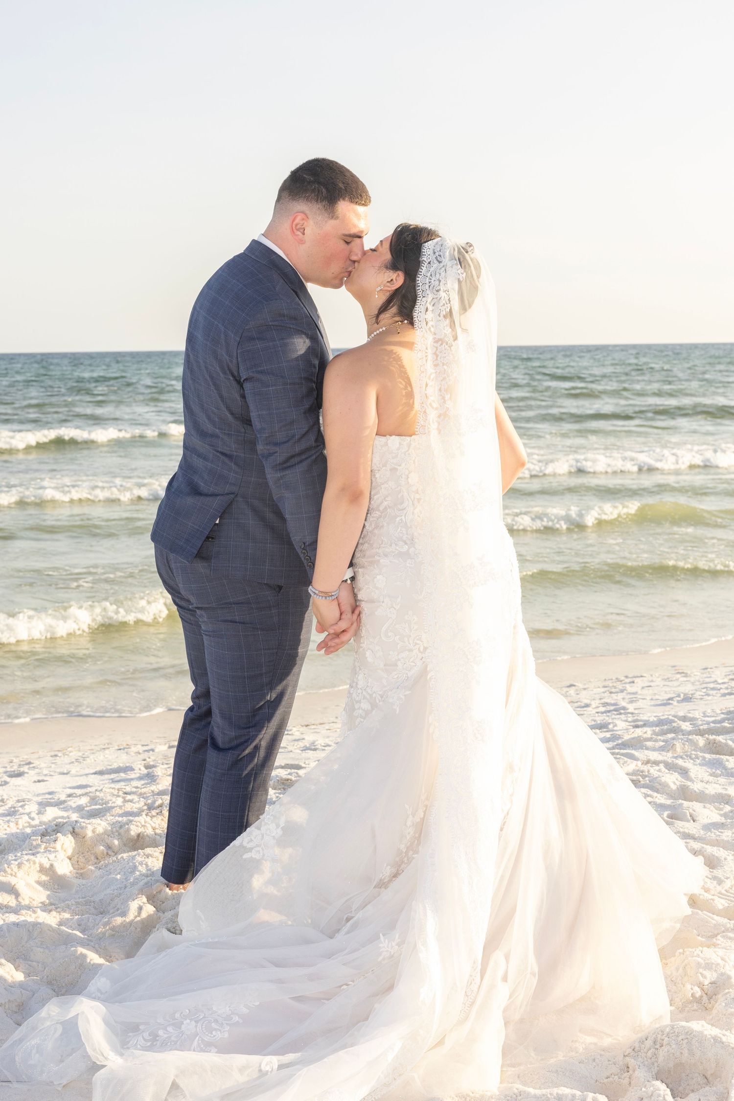 Bride and groom share a romantic kiss on a sandy beach, with ocean waves in the background, bride wearing a lace gown.