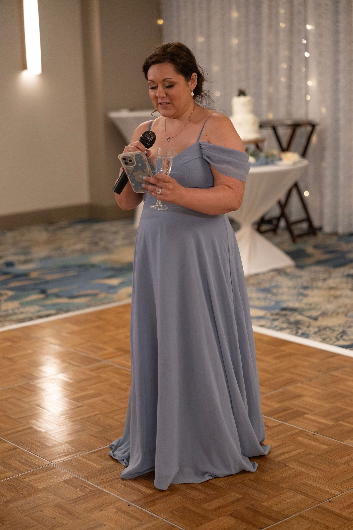 Woman in dusty blue off-shoulder gown giving a toast at a wedding reception, holding microphone and champagne glass.