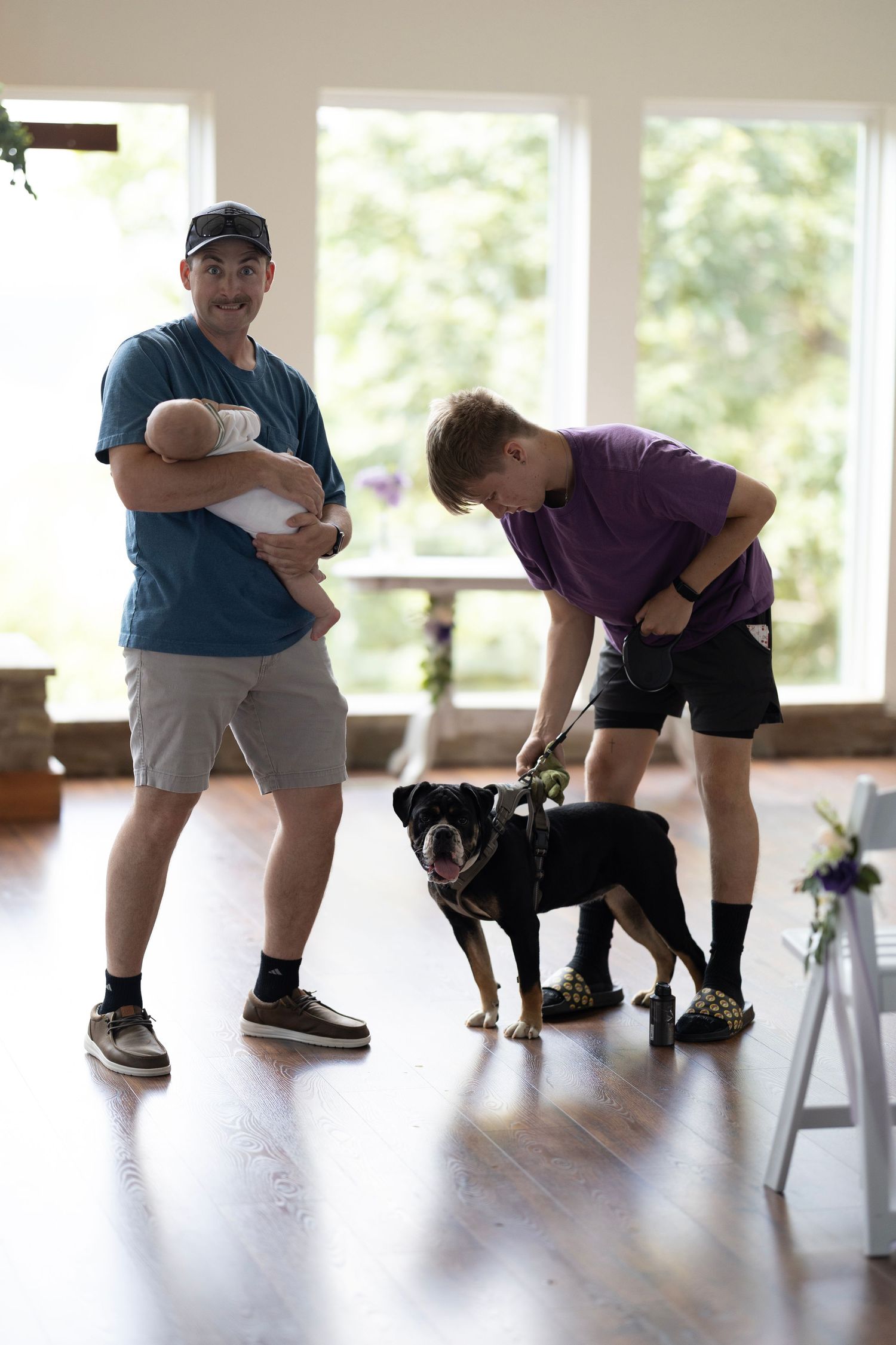 Man holding baby stands beside boy petting a black dog on hardwood floor in bright room.