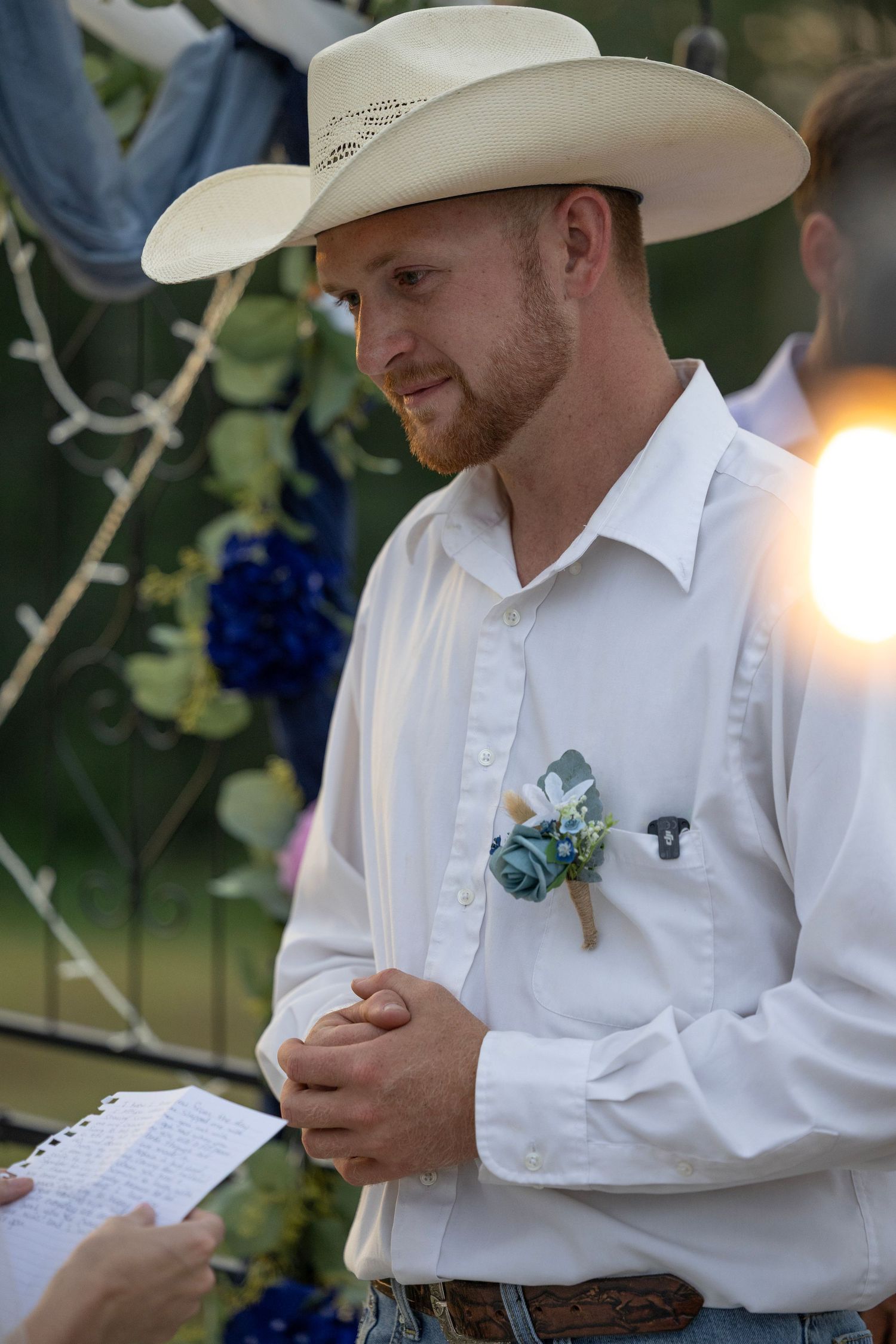 Groom in white cowboy hat and shirt with blue boutonniere reads vows at outdoor western wedding ceremony.