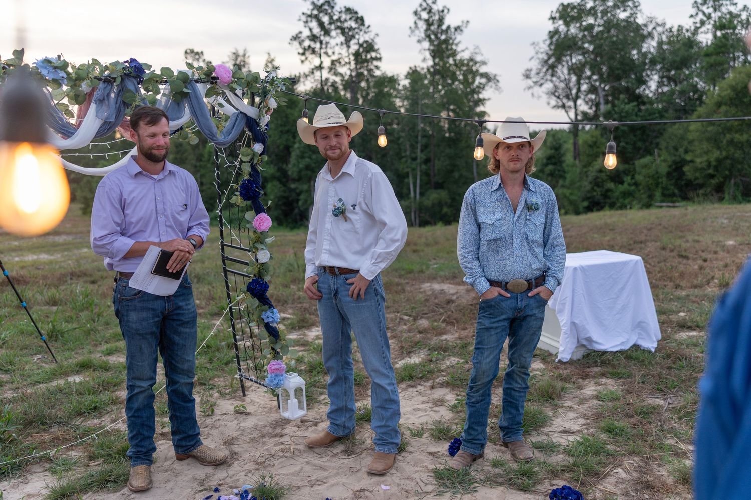 Three people in cowboy hats and jeans stand outdoors at a rustic evening gathering with string lights and floral decor.