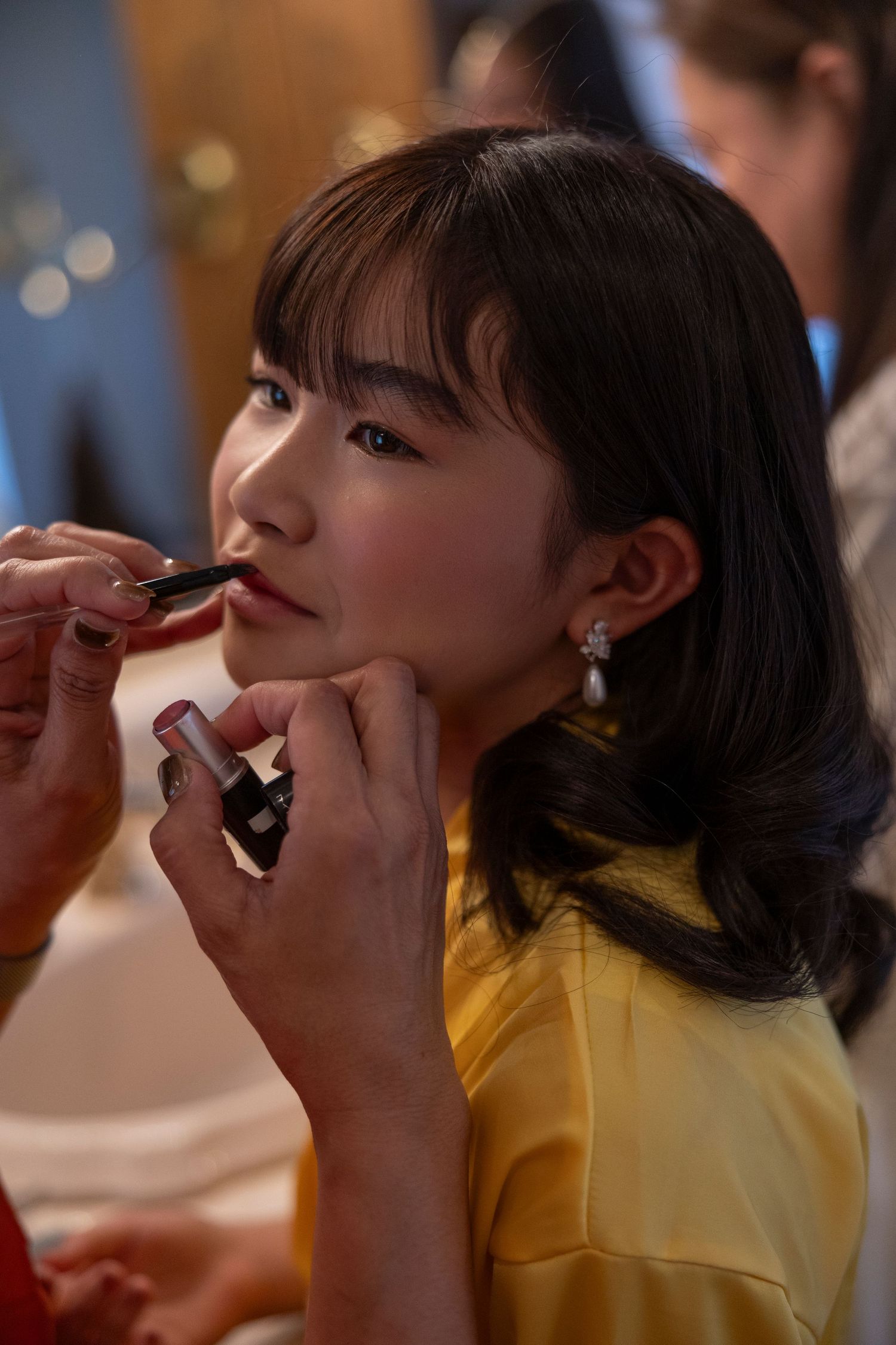A makeup artist applies lipstick to a young Asian woman in a yellow top wearing pearl earrings.
