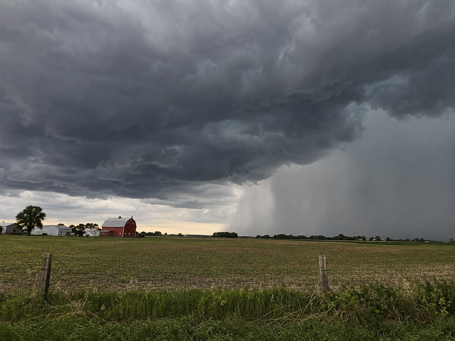 Dark storm clouds loom over a rural farm with a red barn as heavy rain falls across green fields.
