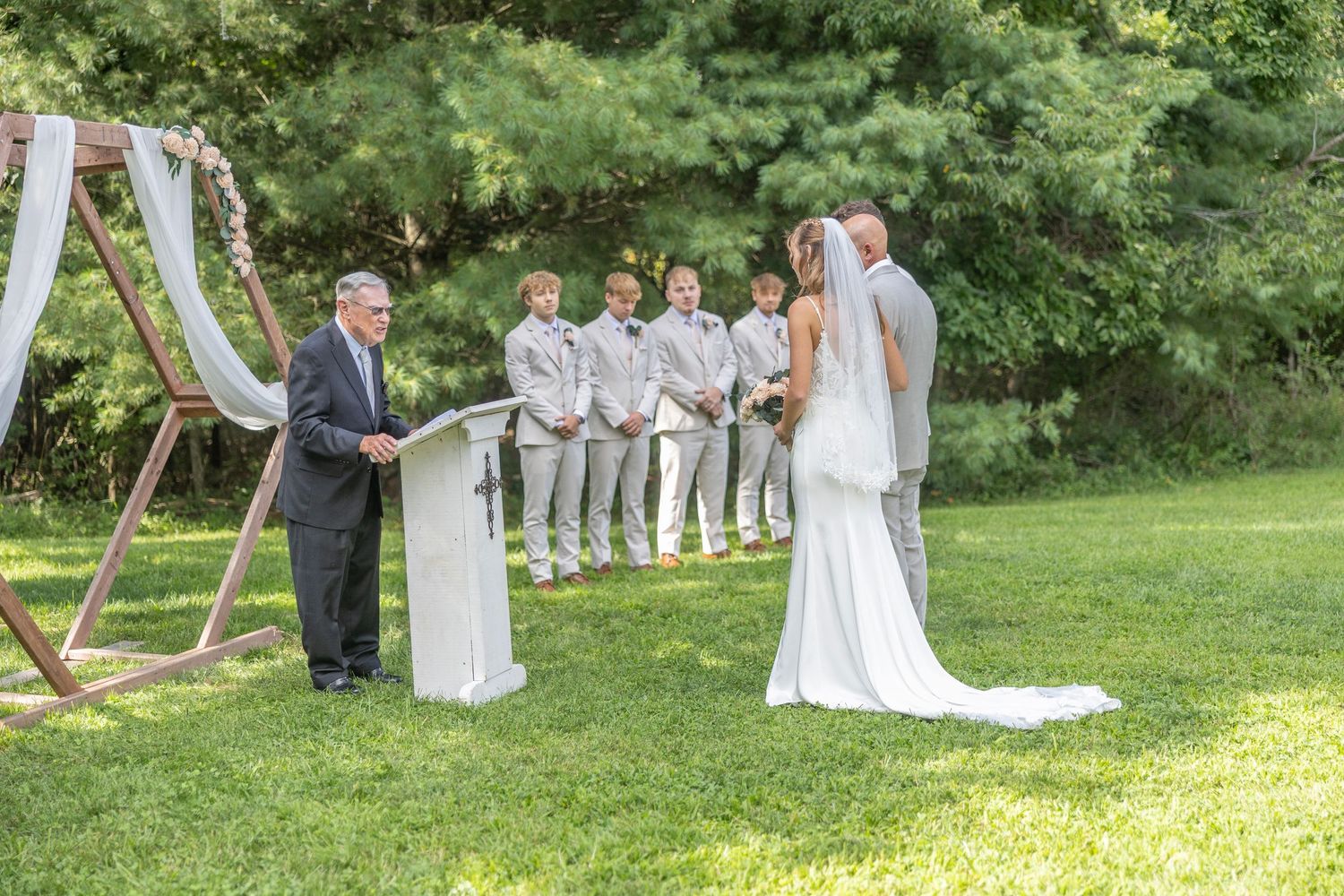 Wedding ceremony setup in a lush garden with white wooden chairs and decorative arches on a sunny day.