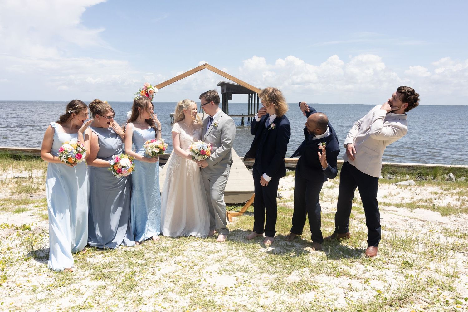 Wedding party in light blue and black attire laughing together on a sunny beach with ocean backdrop.