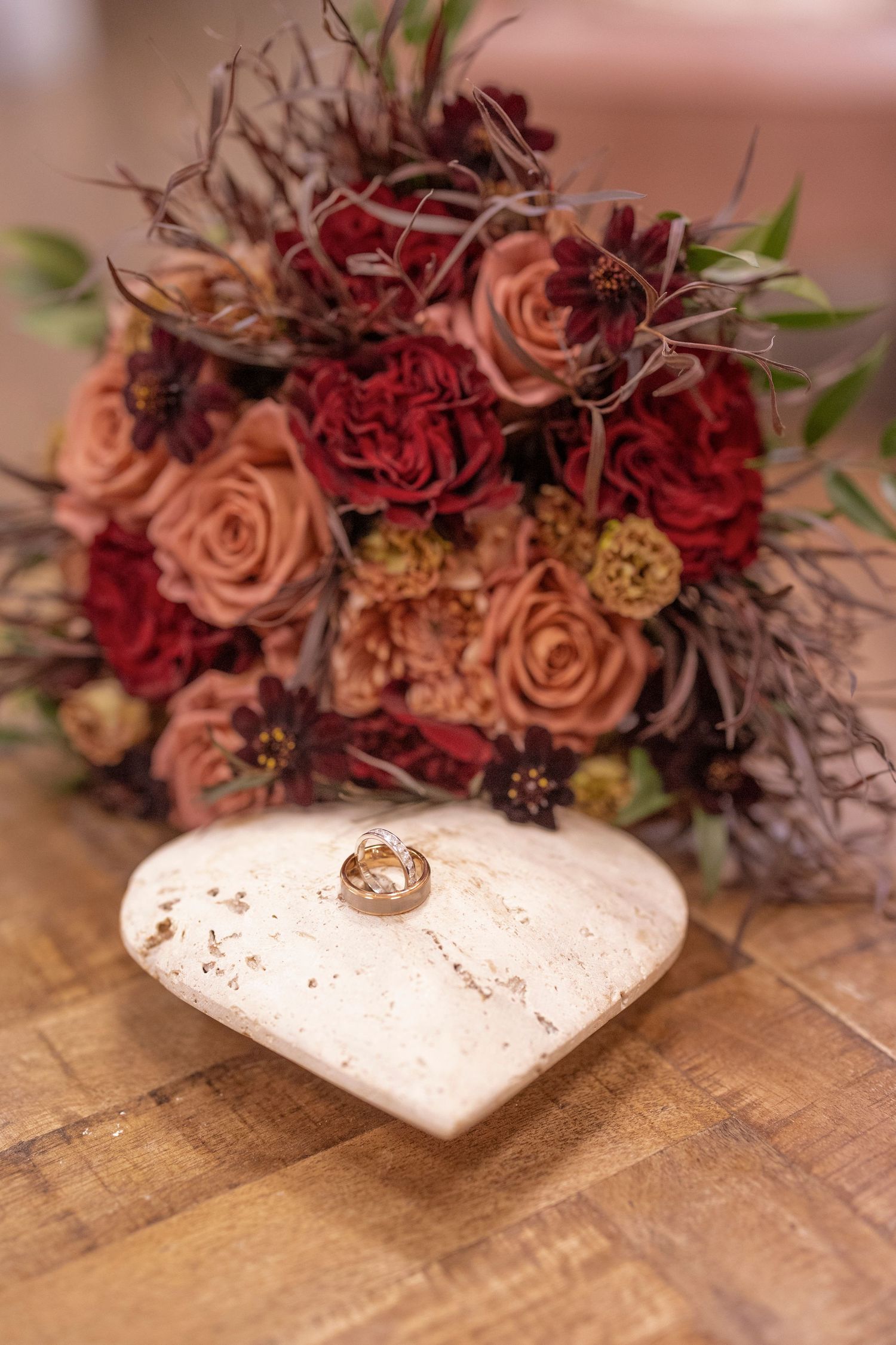 Wedding rings displayed on a white heart-shaped cushion with a romantic autumn bouquet of roses in the background.