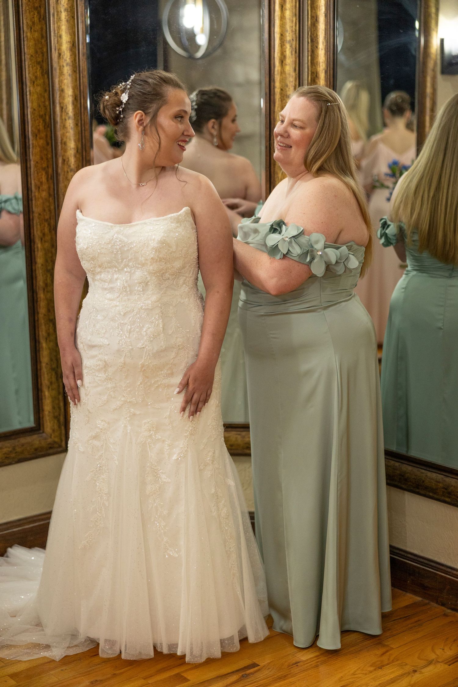 Bride in white lace wedding dress and bridesmaids in sage green dresses standing together in an elegant room with mirrors.